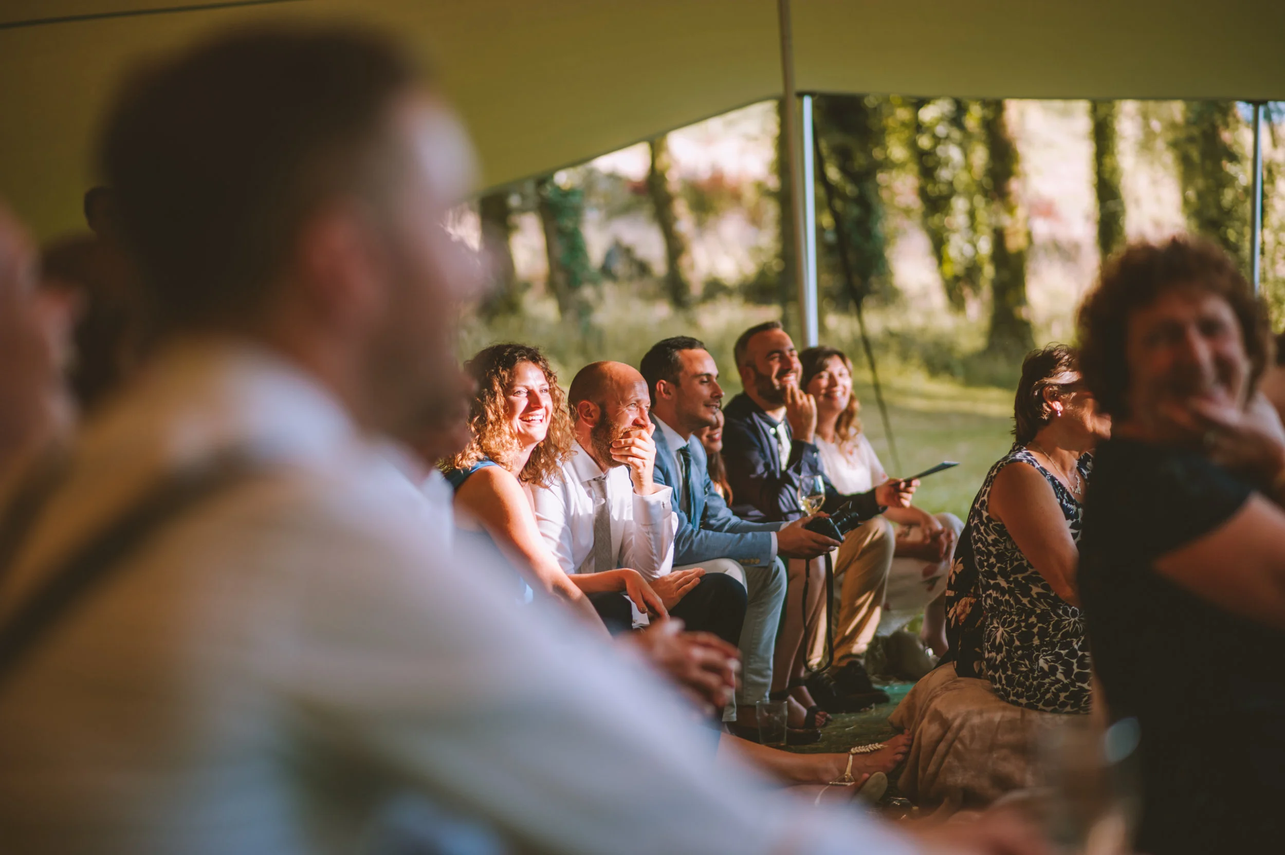 Group of people sitting under a tent outdoors, laughing and smiling during daytime event.