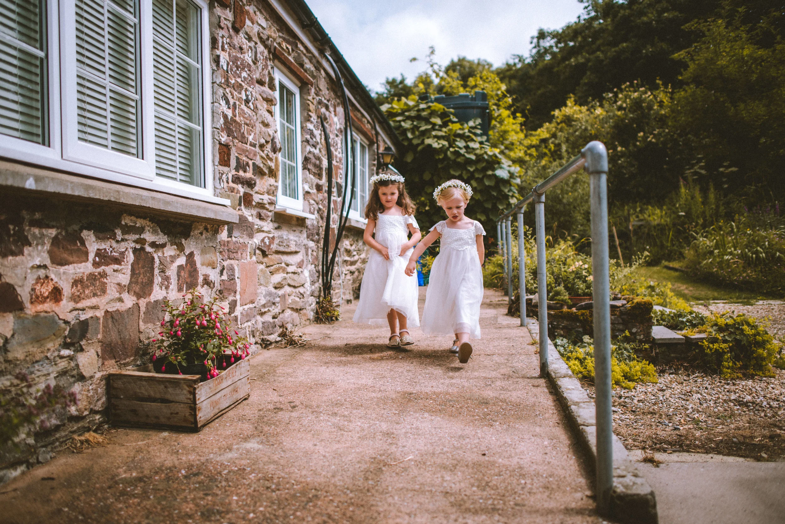 Two young girls in white dresses and floral crowns walking down a concrete pathway beside a stone house with white window frames, surrounded by greenery and a flower bed.