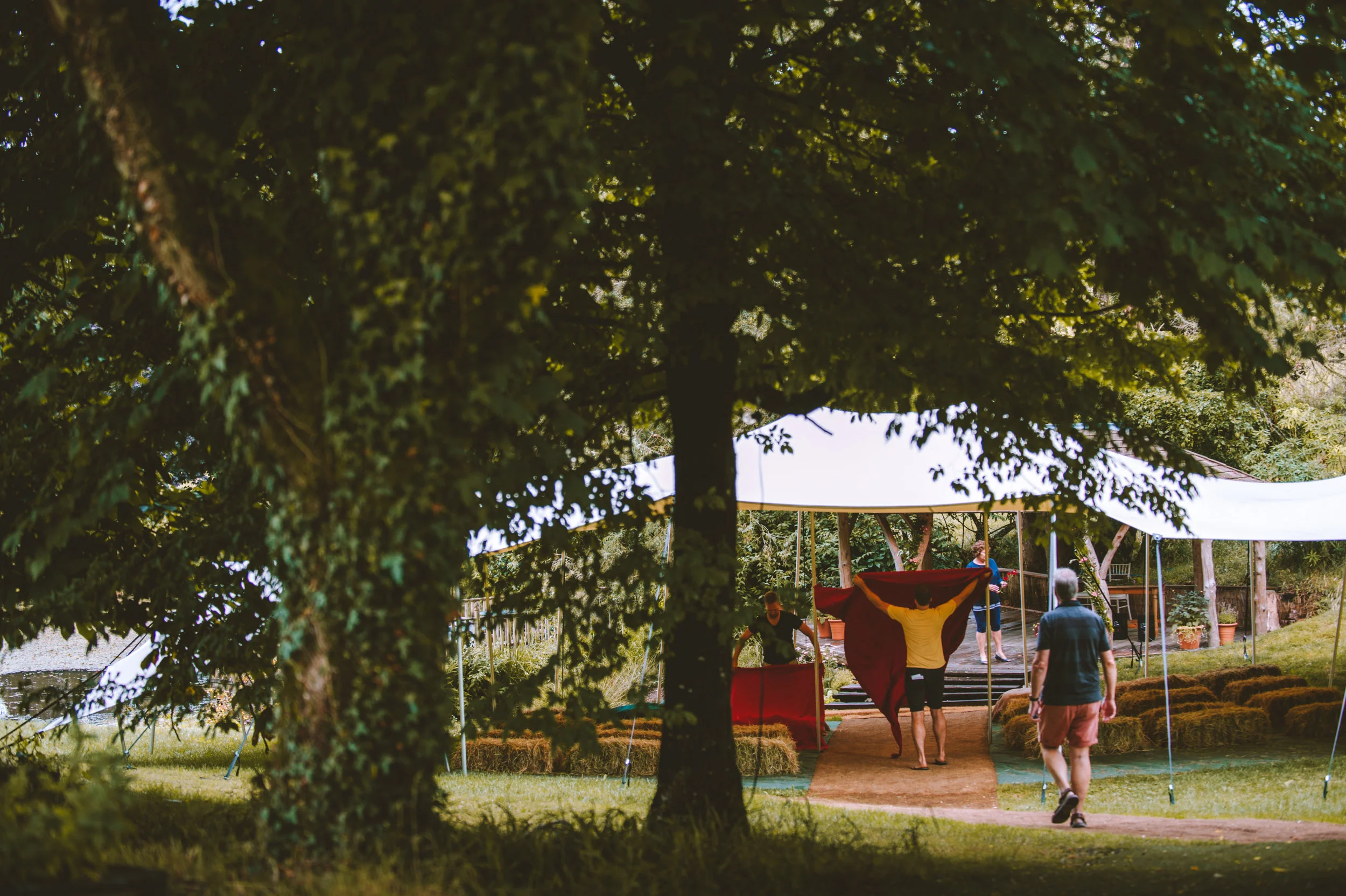 People setting up an outdoor event under a white canopy tent in a lush green park with trees and grass.