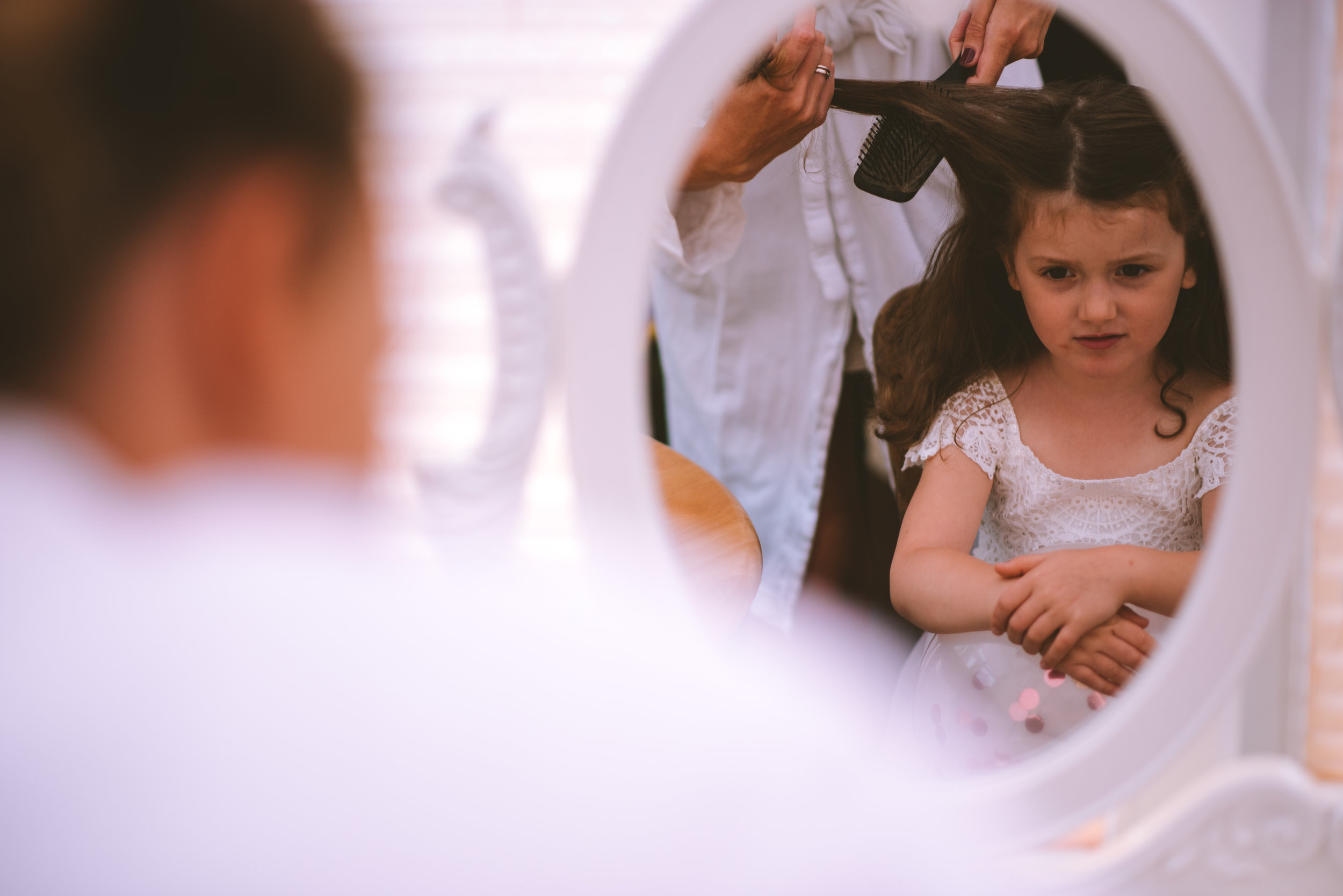 A young girl with long dark hair, wearing a white lace dress, looks at herself in a mirror with a worried expression while getting her hair styled by a person using a hairbrush and comb.