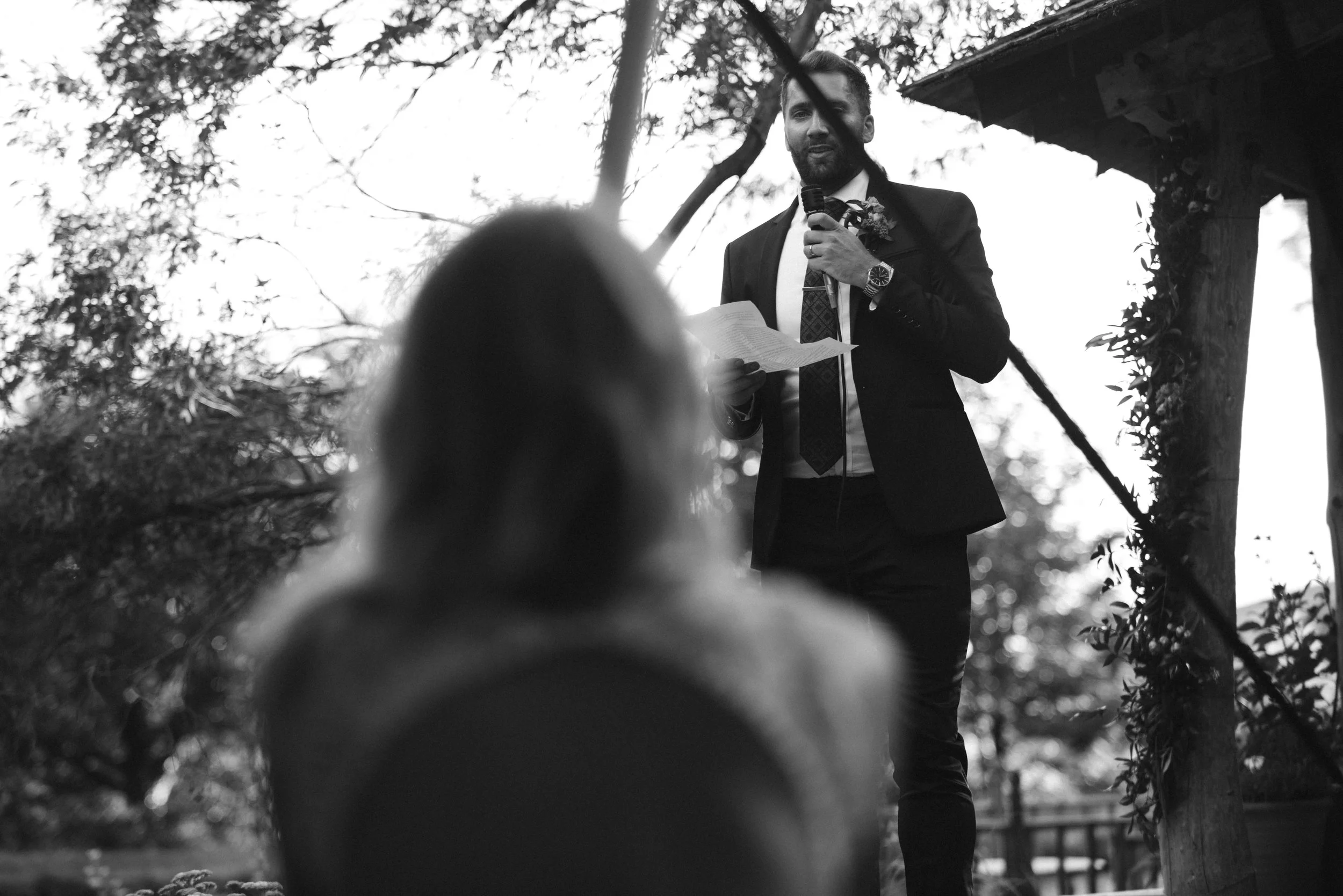 A man in a suit giving a speech at an outdoor wedding ceremony, holding a microphone and reading from a paper, with a blurred person in the foreground.