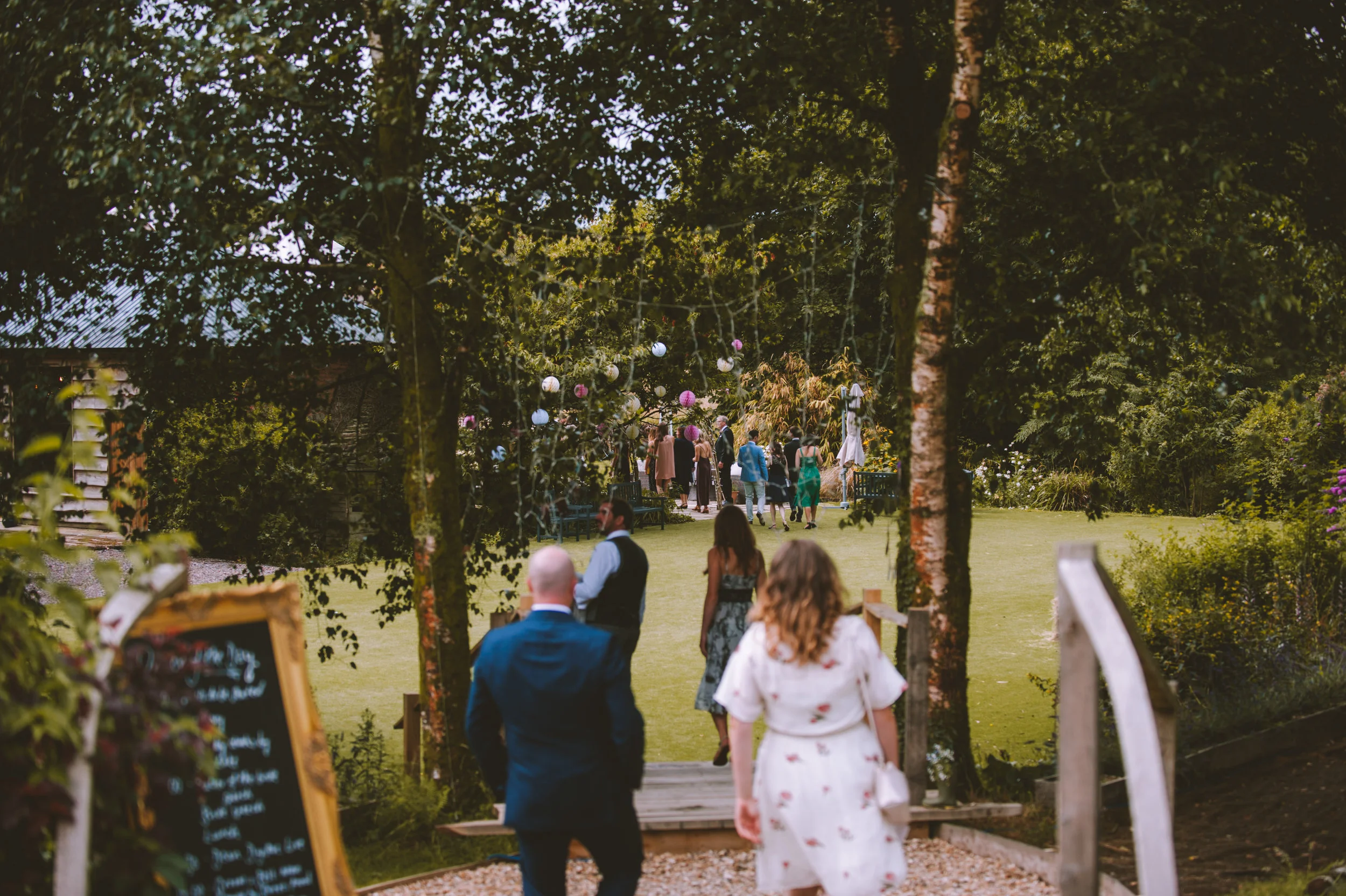 People walking through a garden area decorated with hanging paper lanterns at an outdoor event during the daytime.