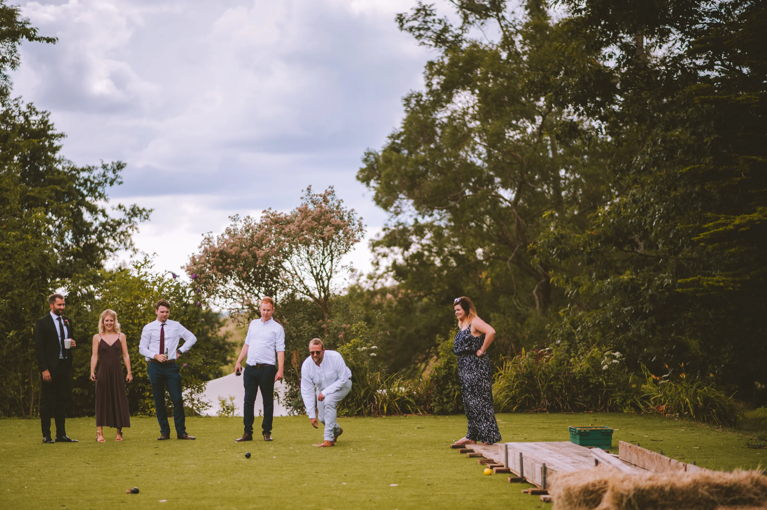 People playing bocce ball outdoors on a grassy field with trees and cloudy sky in the background.