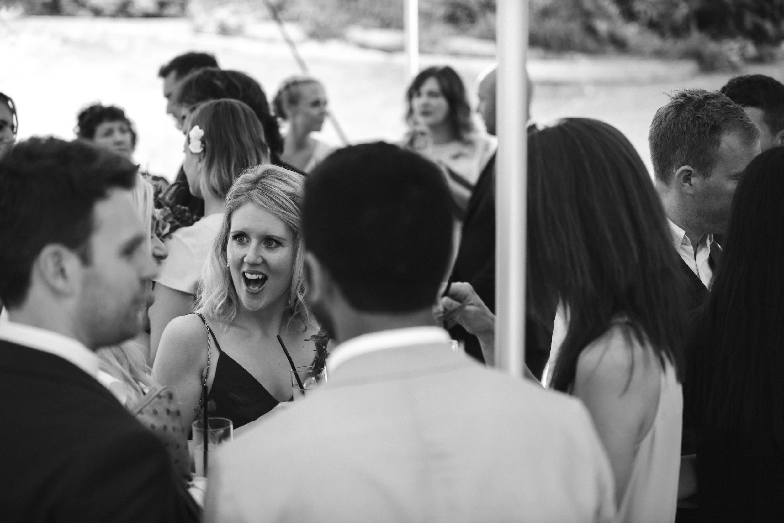 Group of people socializing at an outdoor event, some are talking and drinking, with a woman in the center expressing surprise or excitement.