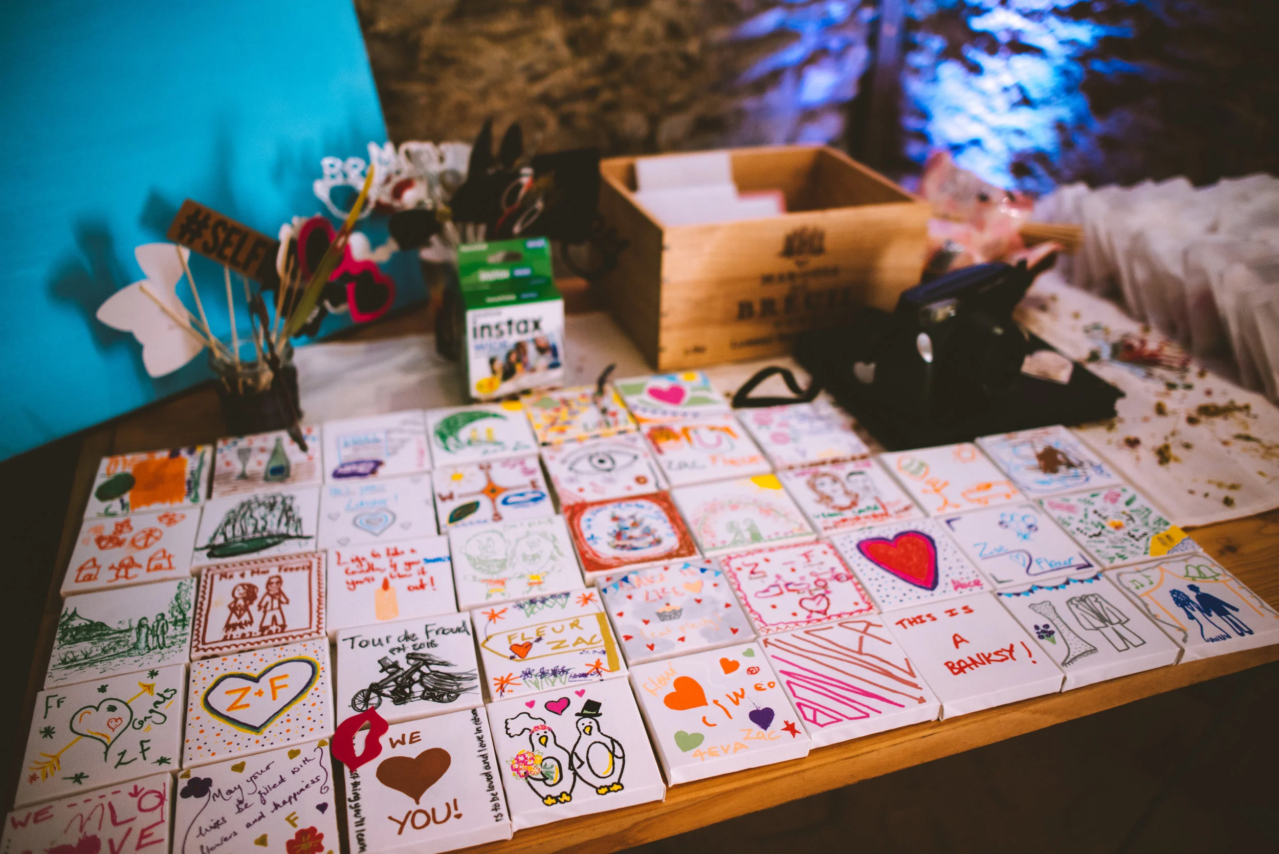 Table with handmade cards decorated with drawings and messages, a box, and photo booth props including black and white masks and signs, set against a stone wall backdrop
