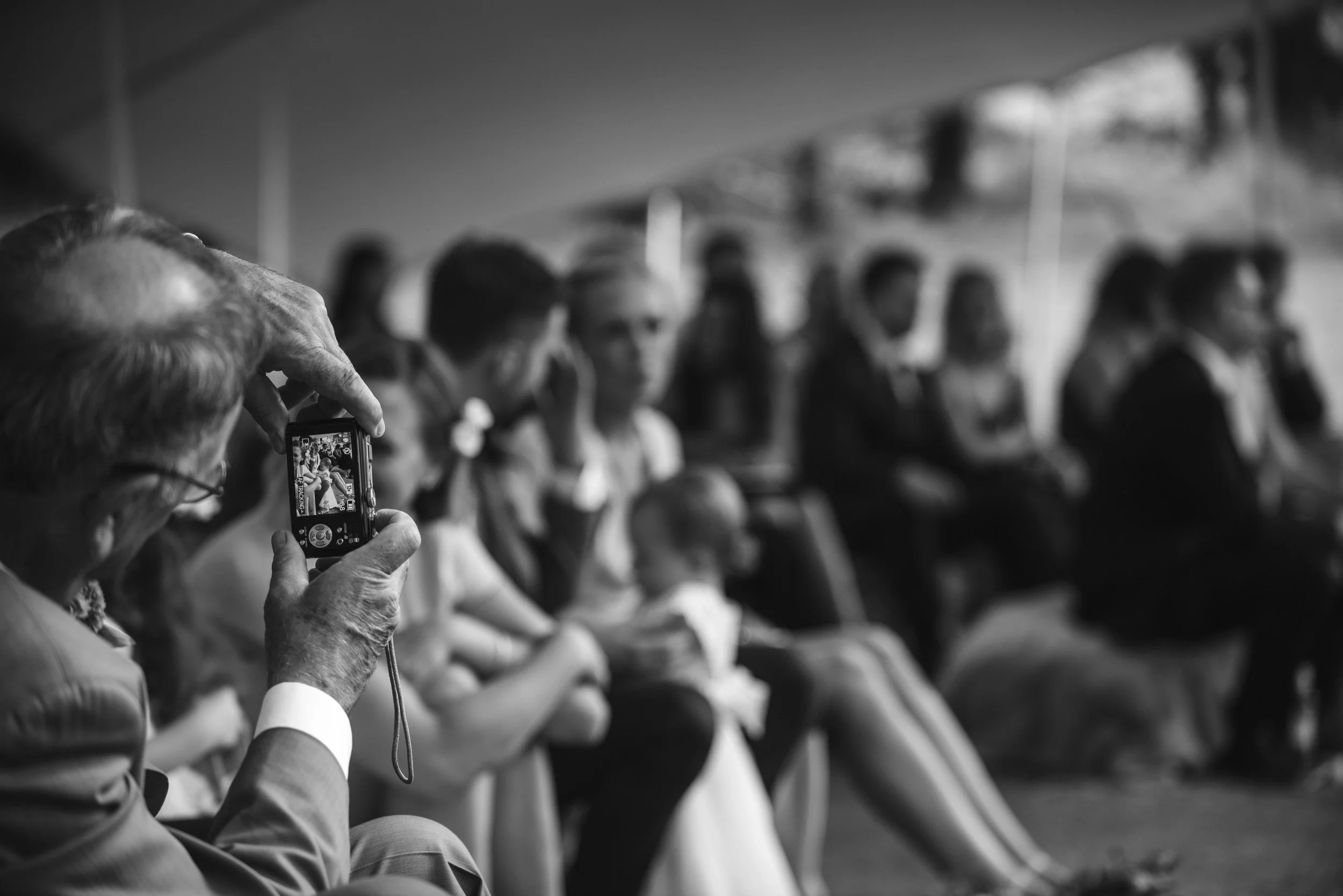 An elderly man taking a photo with a small digital camera of a seated group of people, some of whom are in formal attire, in an indoor setting.
