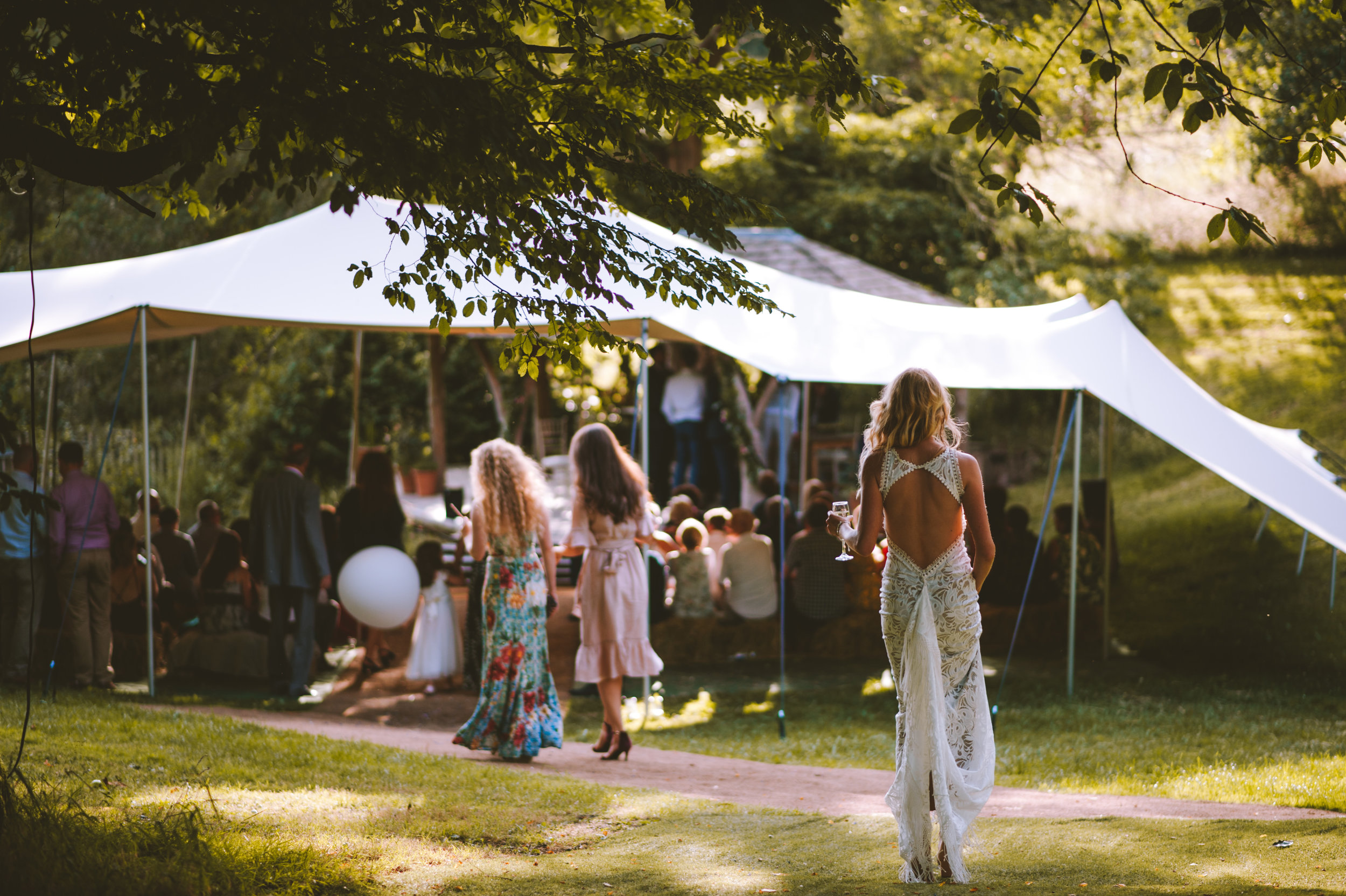 An outdoor wedding reception under a large white canopy with guests seated and standing around. A woman in a white lace dress with an open back and blonde hair is walking away, holding a glass of wine, in the foreground. The scene is surrounded by lu