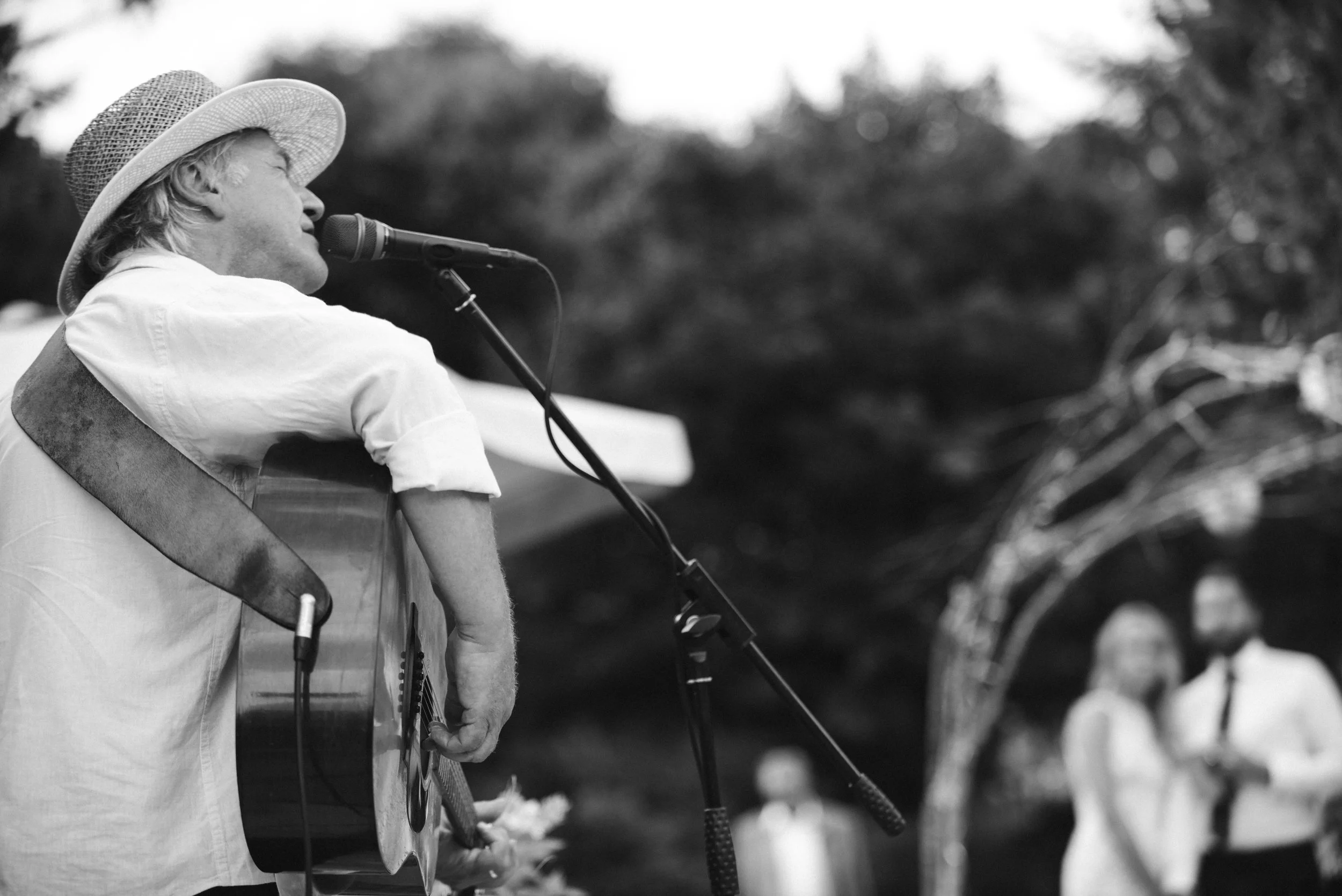 A man in a straw hat playing an acoustic guitar and singing into a microphone at an outdoor event, with blurred audience in background.