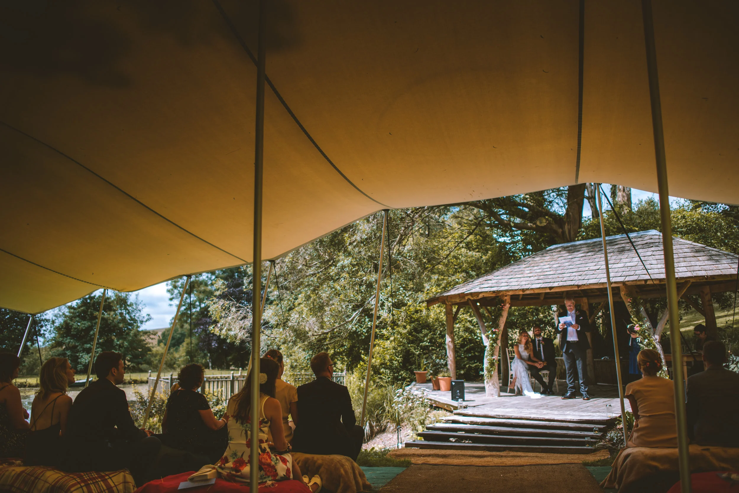 Outdoor wedding ceremony with seated guests under a canopy, a person reading at a small wooden stage, and a rustic gazebo in the background surrounded by trees and greenery.
