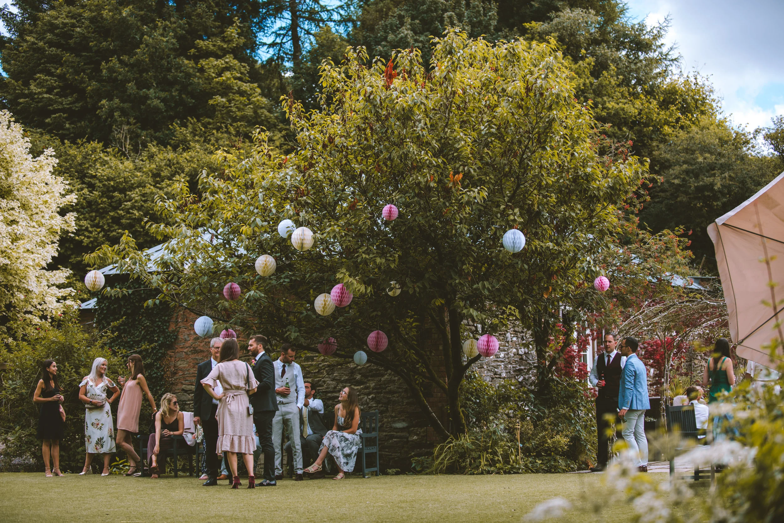 People gathered outdoors at a garden party, standing and sitting on benches under a tree decorated with colorful paper lanterns, with lush green trees in the background.