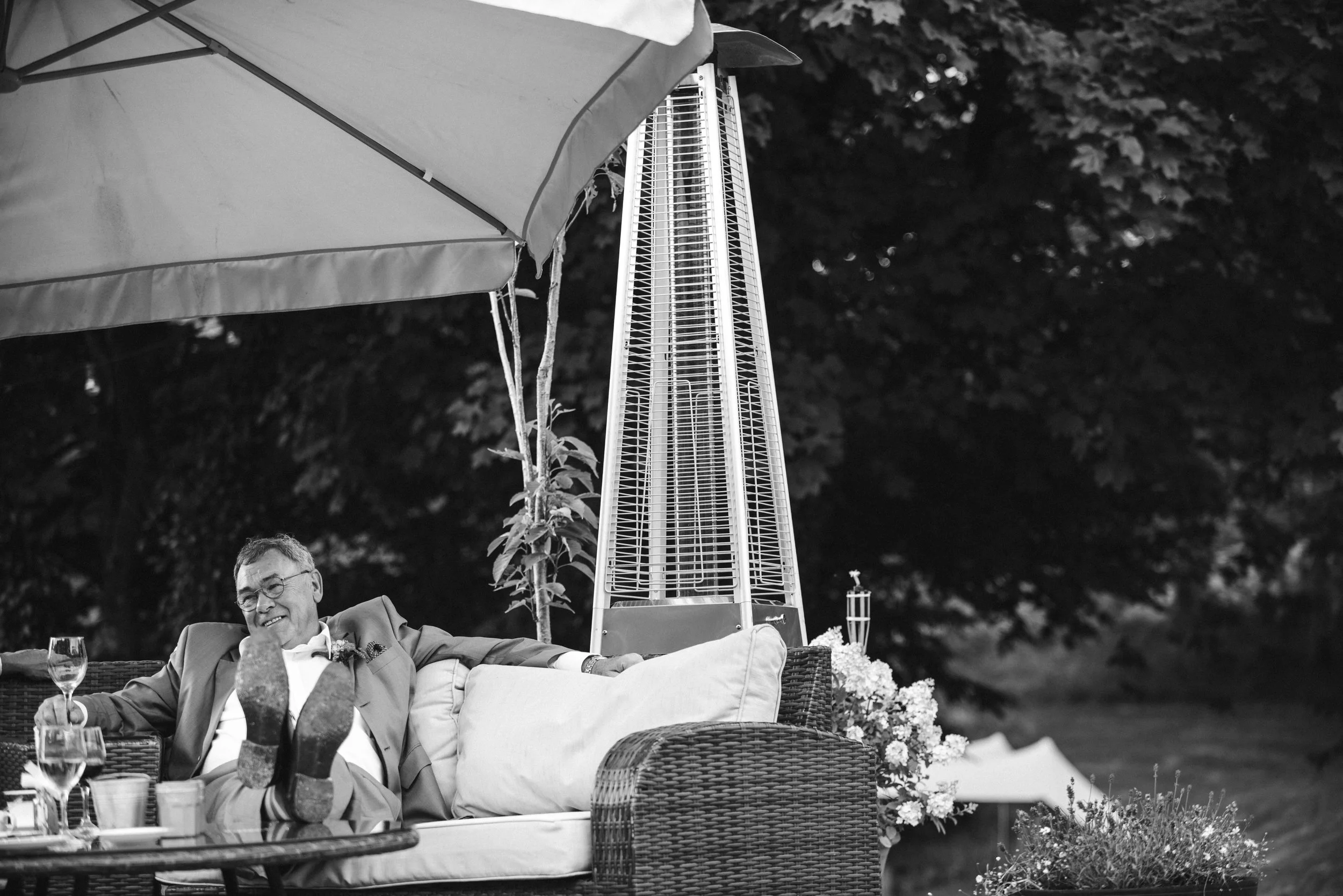 A man in a suit relaxing and smiling on a wicker outdoor sofa, with his feet up on the table, drinks and glassware in front of him, under a large patio umbrella, outdoors with trees in the background.
