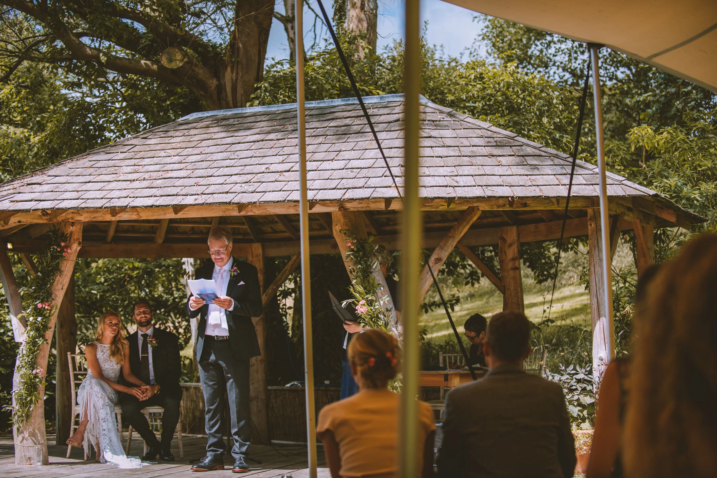 A wedding ceremony outdoors under a wooden gazebo, with a man reading vows or a speech, a bride and groom sitting nearby, and guests seated in front watching.