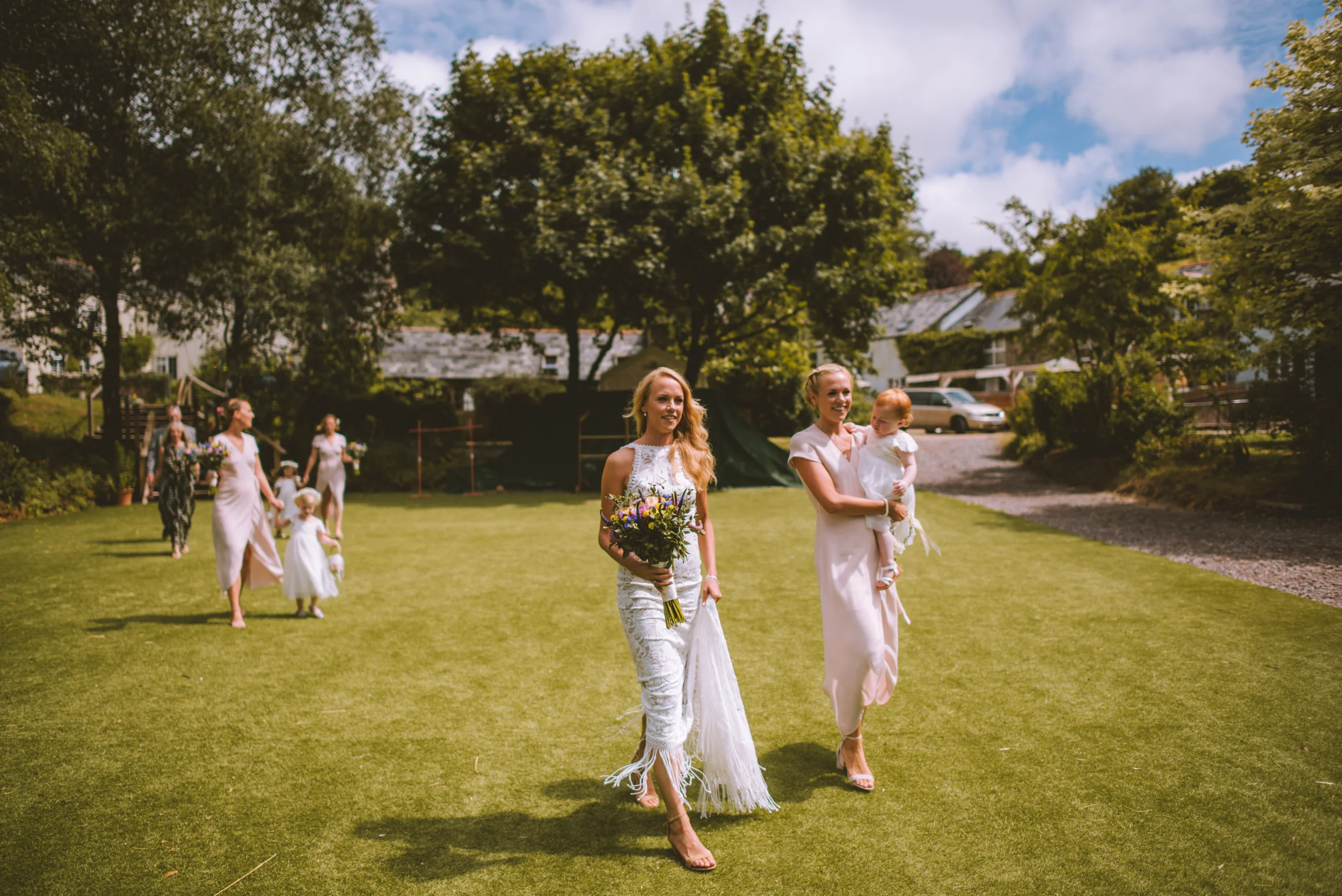 Women in white dresses walking on a grassy lawn, holding flowers, with trees and houses in the background.