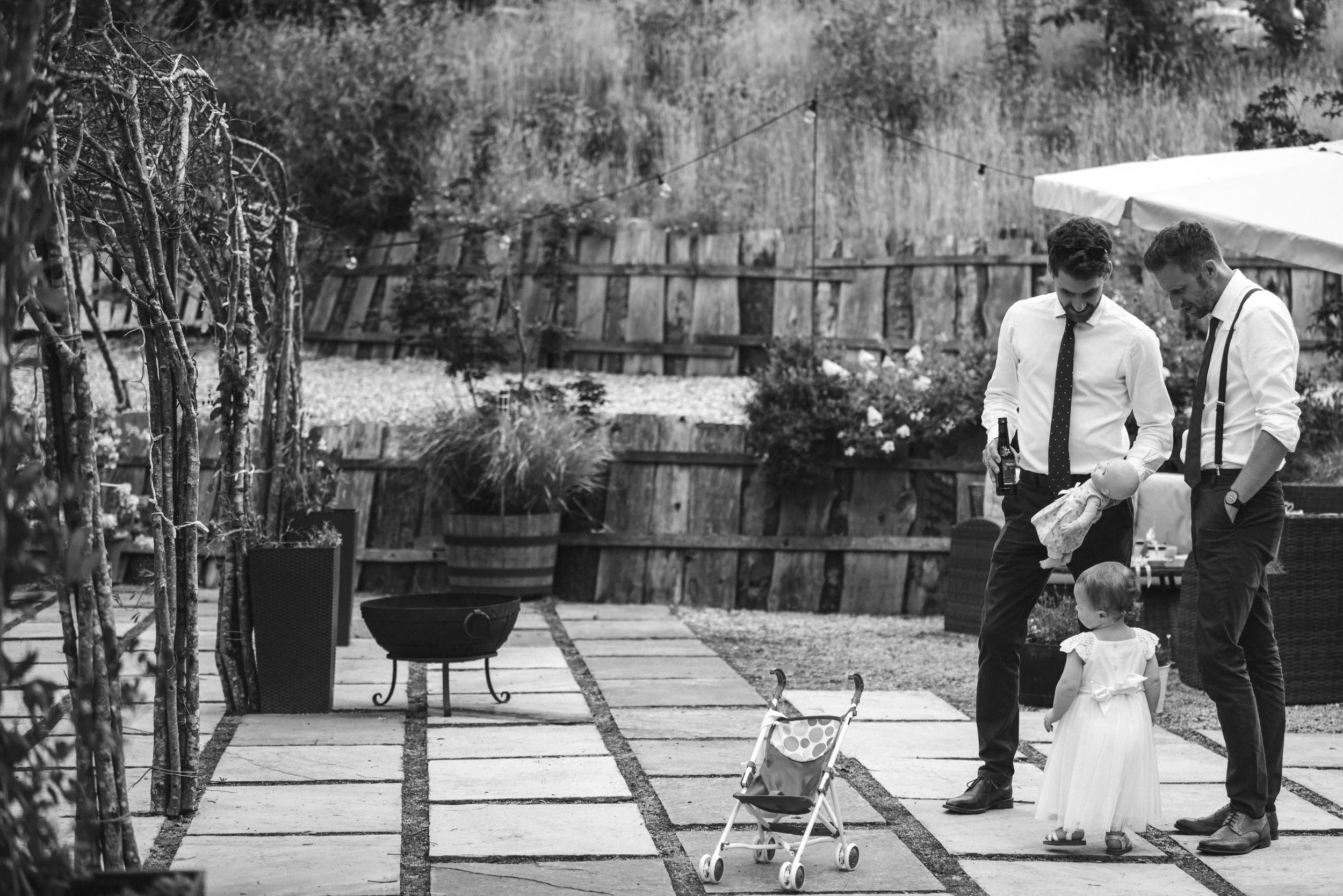 Two men in dress shirts and ties playing with children in a backyard garden on a paved walkway, with a stroller nearby. One man holds a bottle, and a toddler girl in a white dress stands looking at them. The garden has potted plants, a wooden fence, 