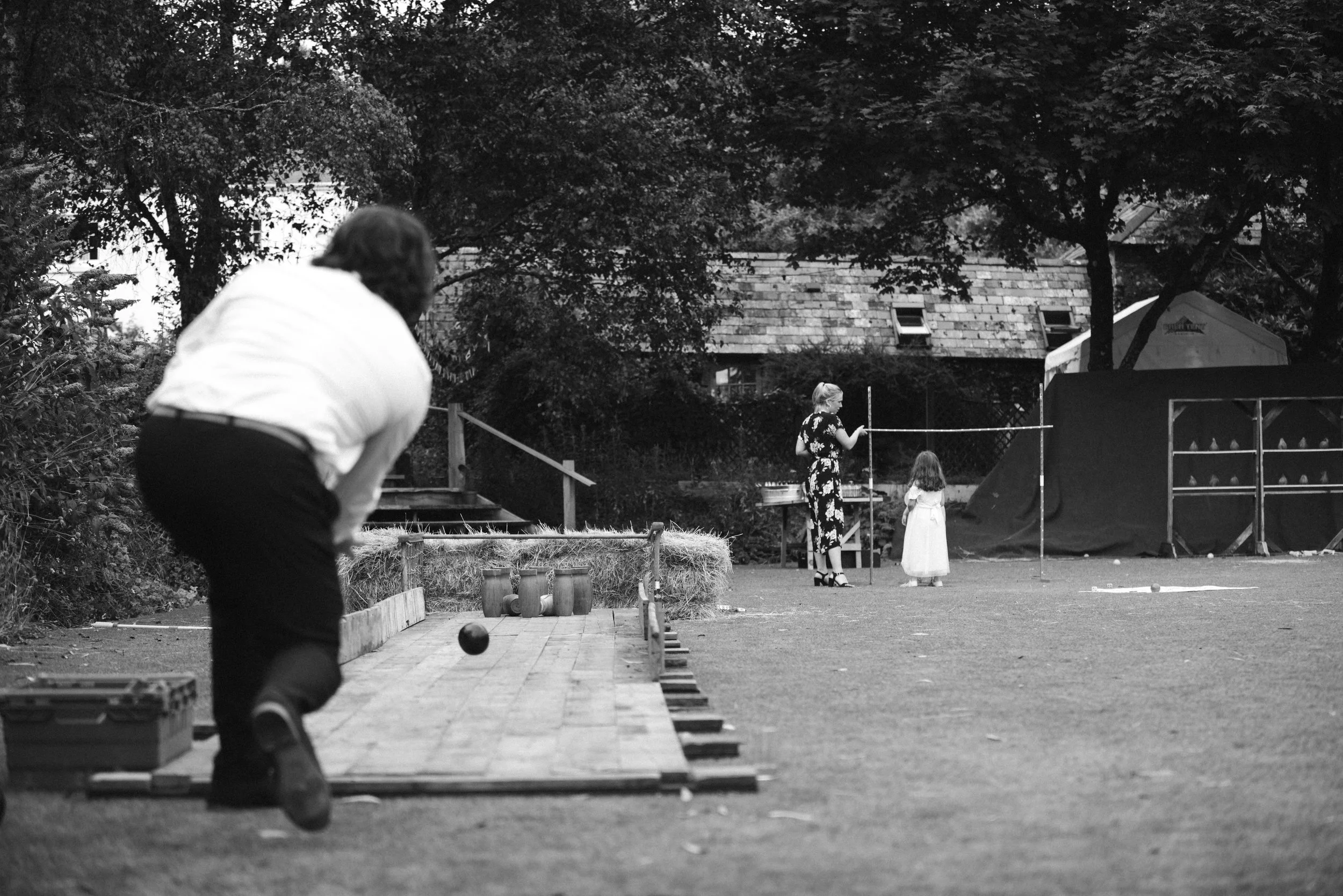A person in the foreground is playing lawn bocce, with three bocce balls and a box nearby. In the background, a woman and a young girl are standing by a string line, possibly playing badminton or a similar game, on a grassy area with trees, a house, 