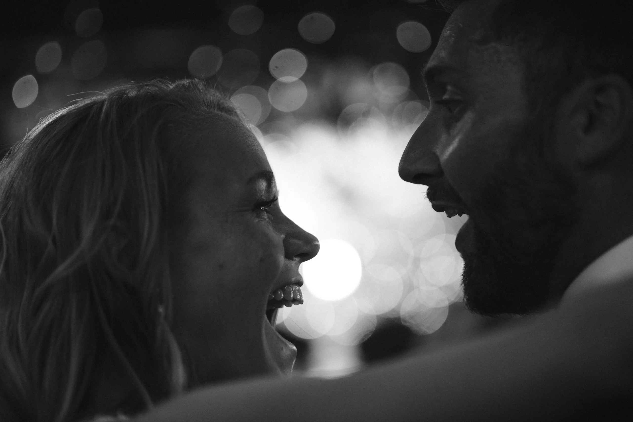 A close-up black and white photo of a man and woman facing each other, smiling, with bokeh lights in the background.