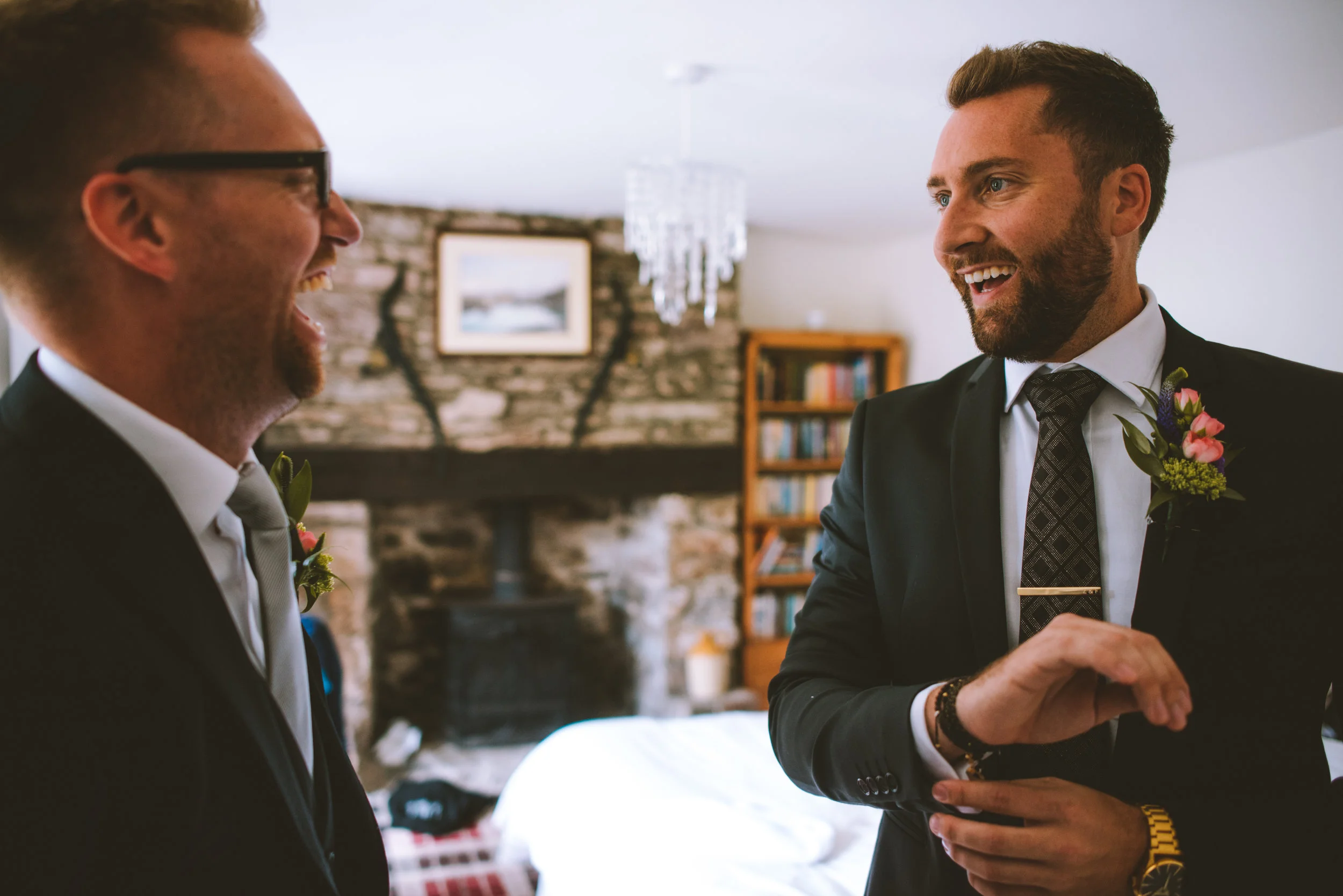 Two men in suits with boutonnieres smiling and talking indoors, with a fireplace and bookshelf in the background.