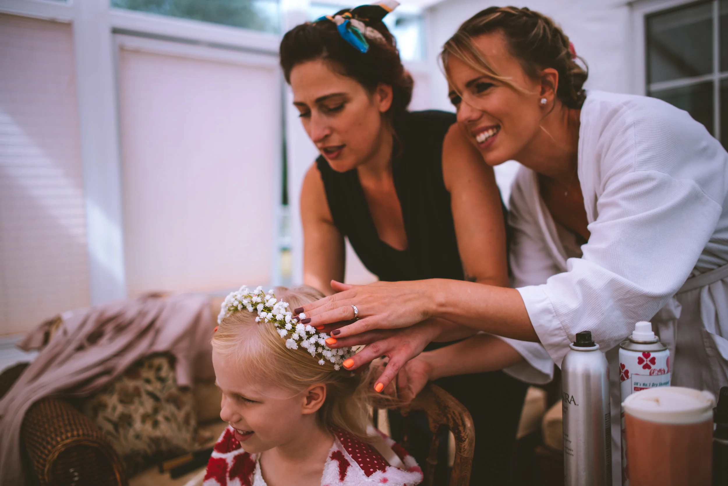 Two women dress a young girl with a floral headband, possibly preparing her for a special event. The women are smiling and look happy.