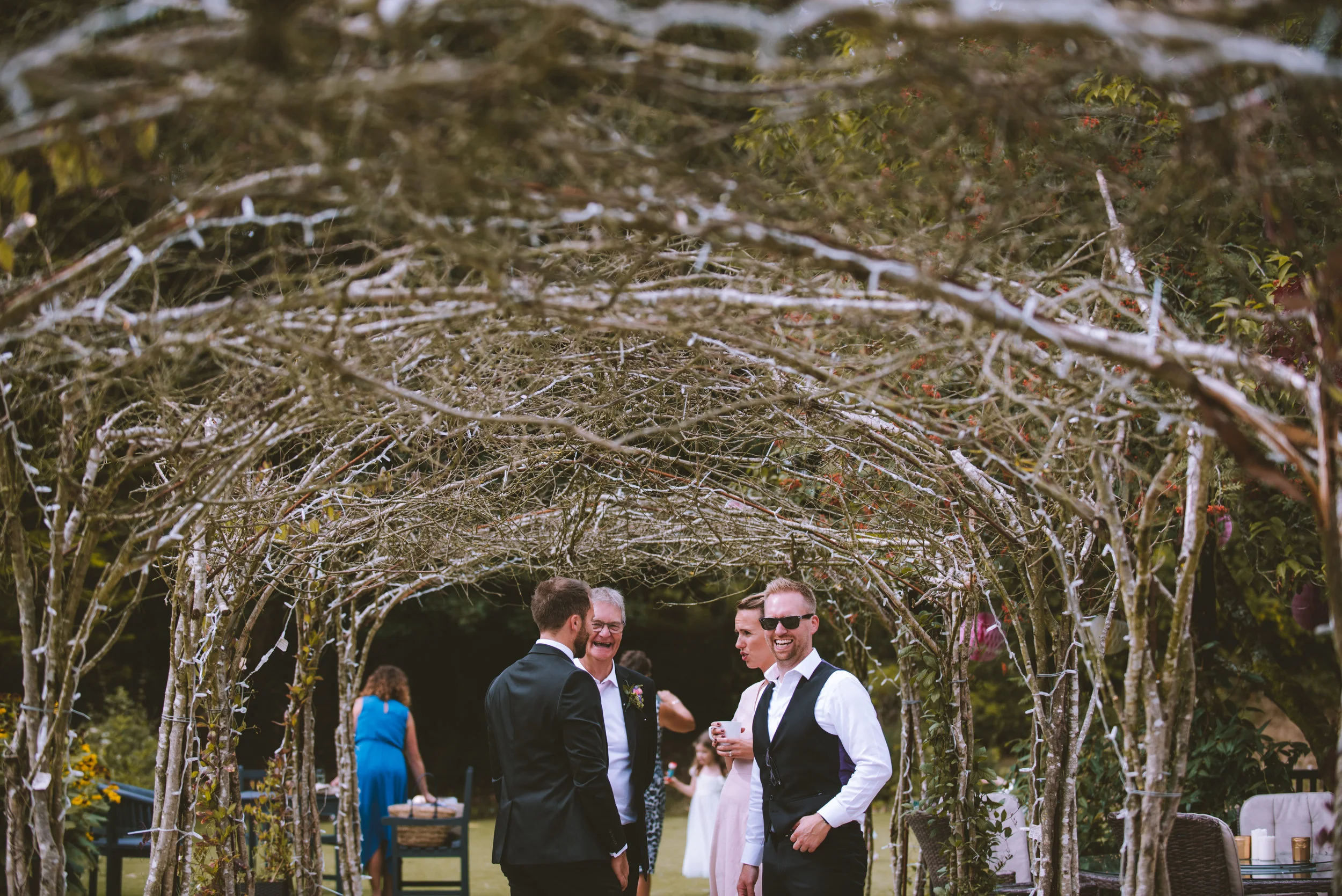 People at a wedding or outdoor event sharing smiles and conversation under a decorative arched garden structure.
