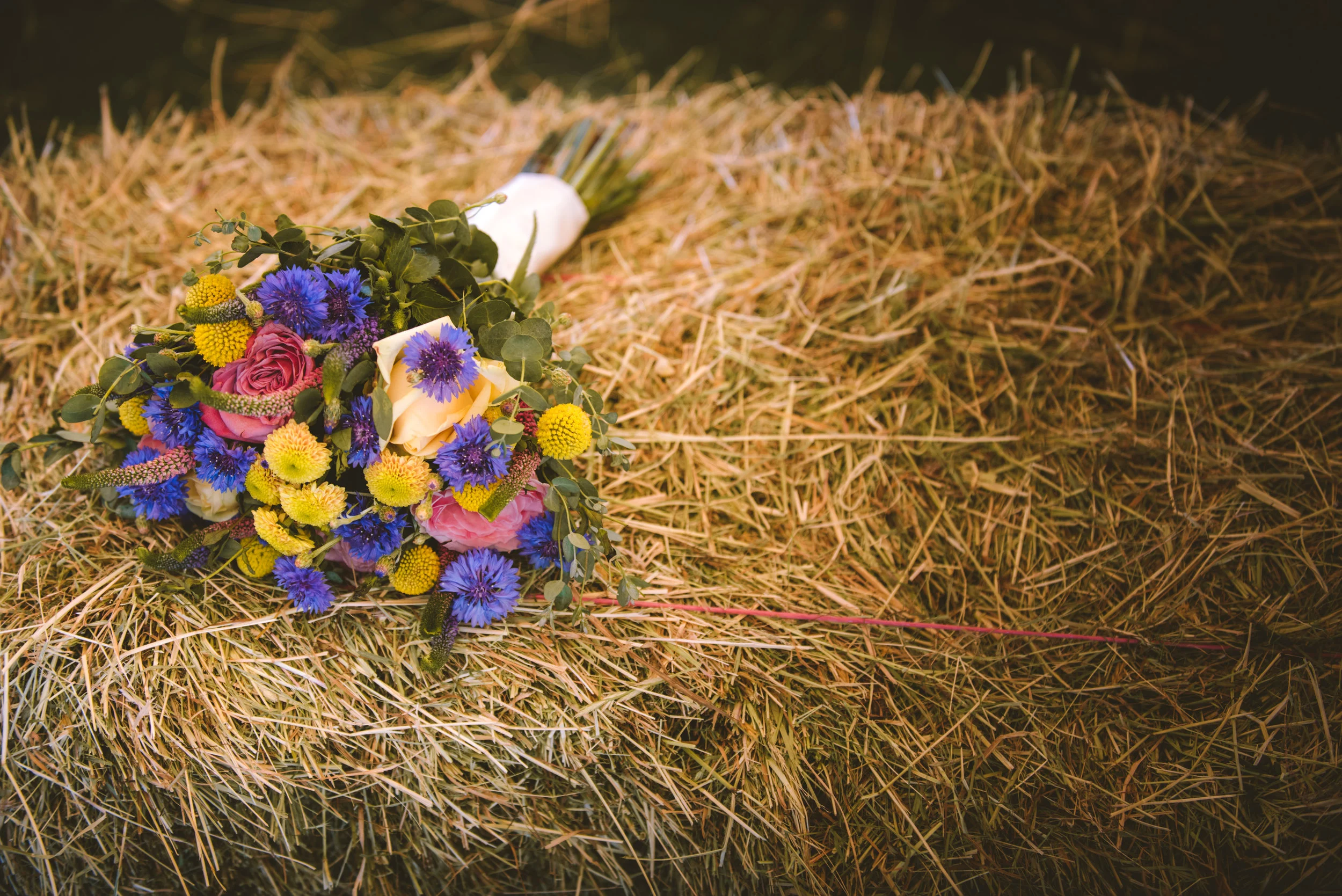 A bouquet of colorful flowers including pink roses, yellow billy balls, and purple cornflowers, resting on a bed of hay.