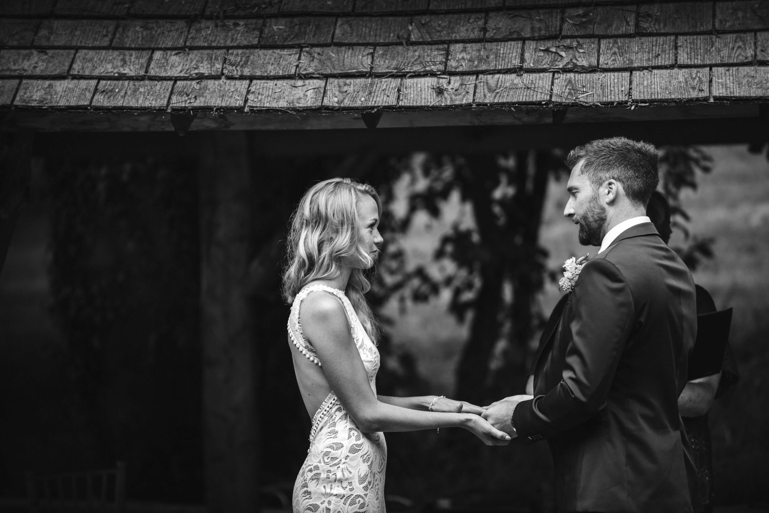 A bride and groom holding hands during their wedding ceremony outdoors, with the bride in a lace dress and the groom in a suit, under a roof with trees in the background.