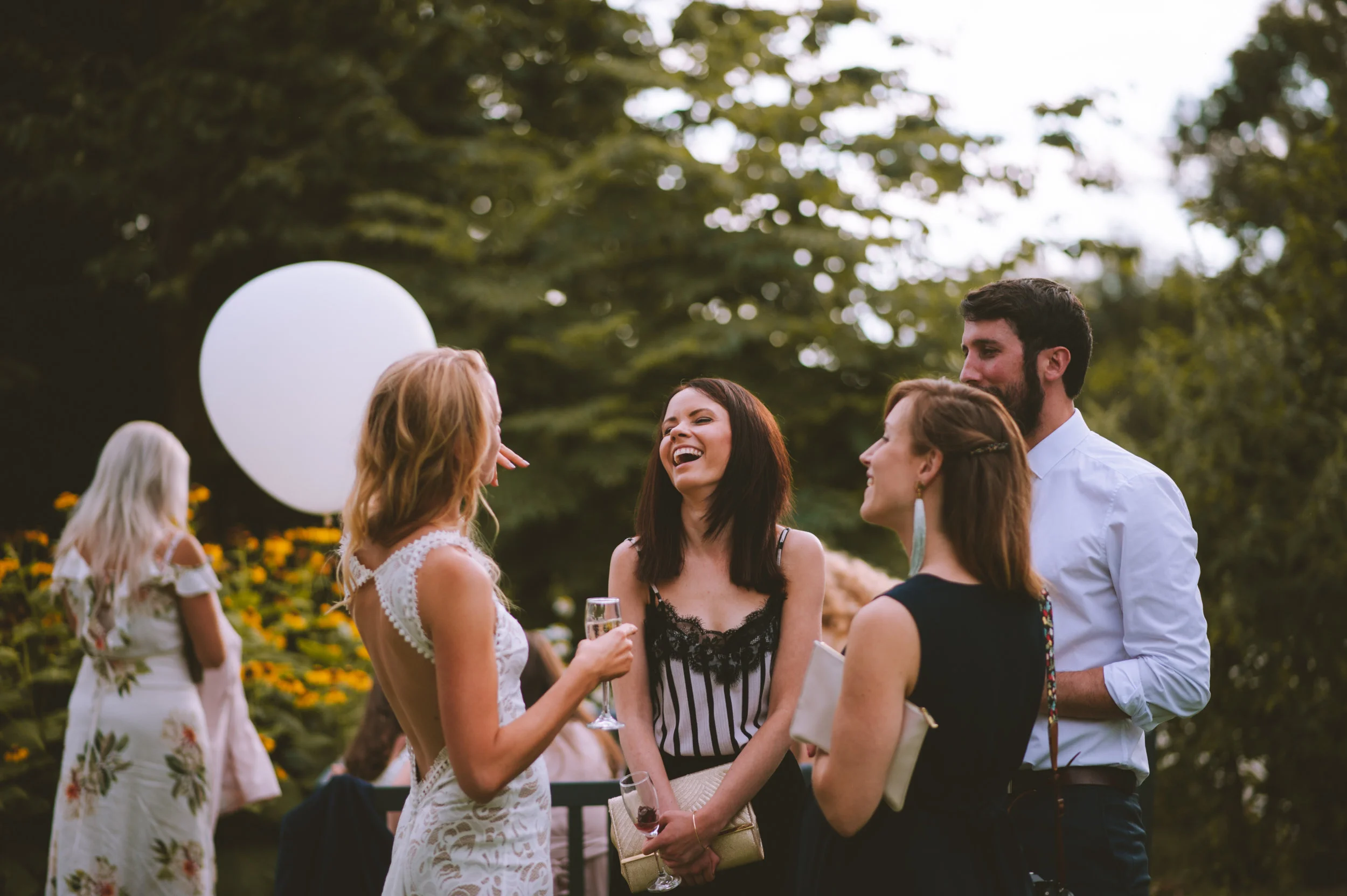 Group of five friends at an outdoor gathering, with four in a conversation in the foreground and one in the background, with a tree and flowers behind them.