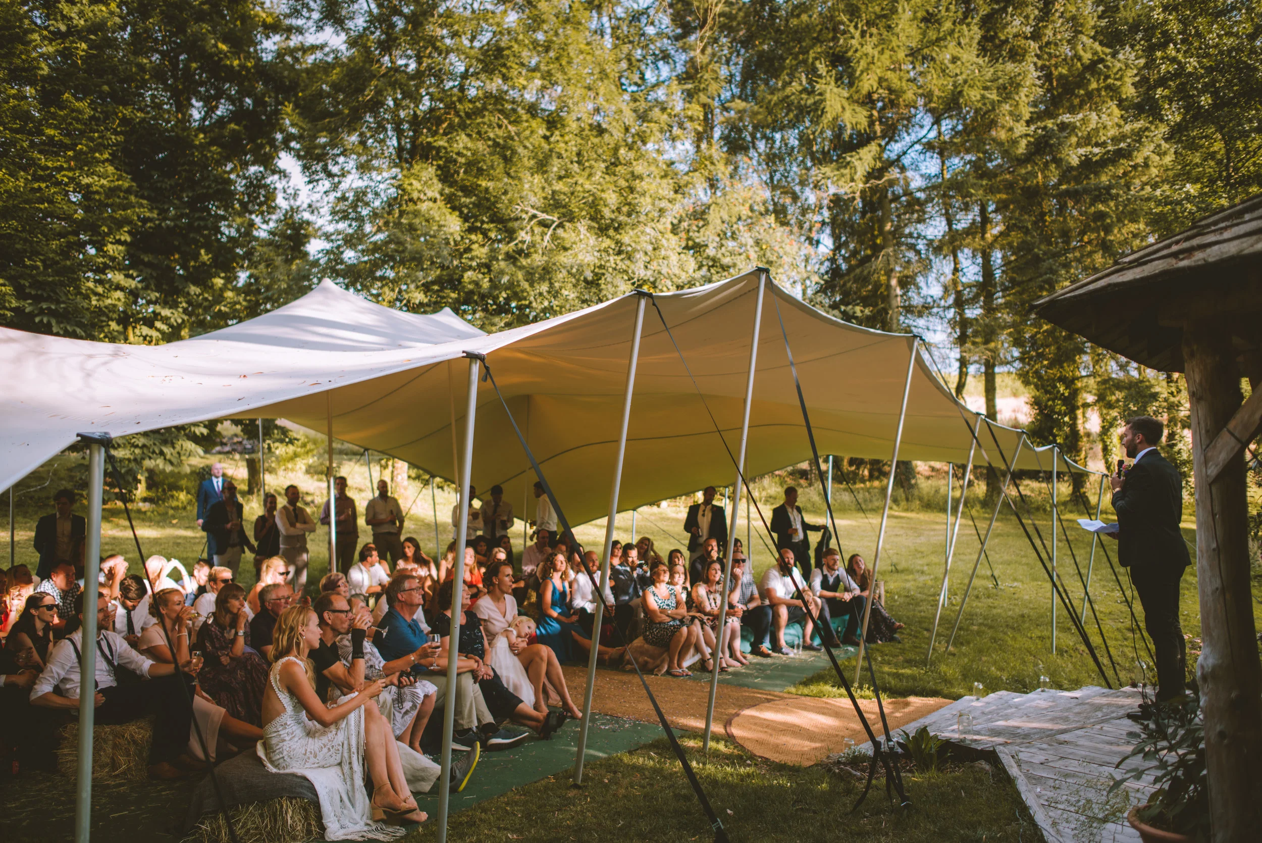 People attending an outdoor wedding under a white canopy, listening to a man in a suit giving a speech, surrounded by tall trees and green grass.