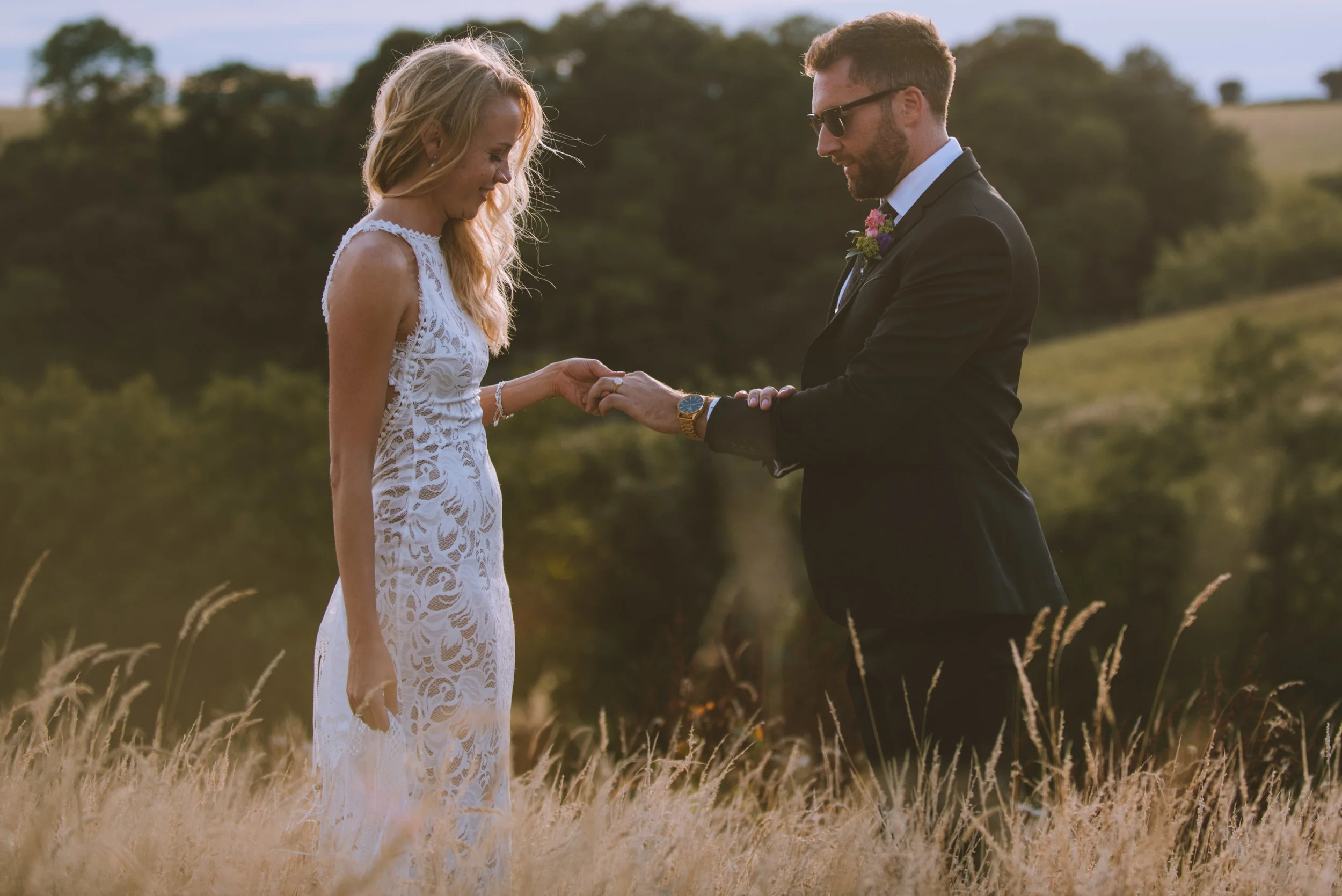 A couple during a wedding ceremony in an outdoor field, holding hands and looking at each other. The woman is wearing a white lace dress and the man is in a black suit with sunglasses.