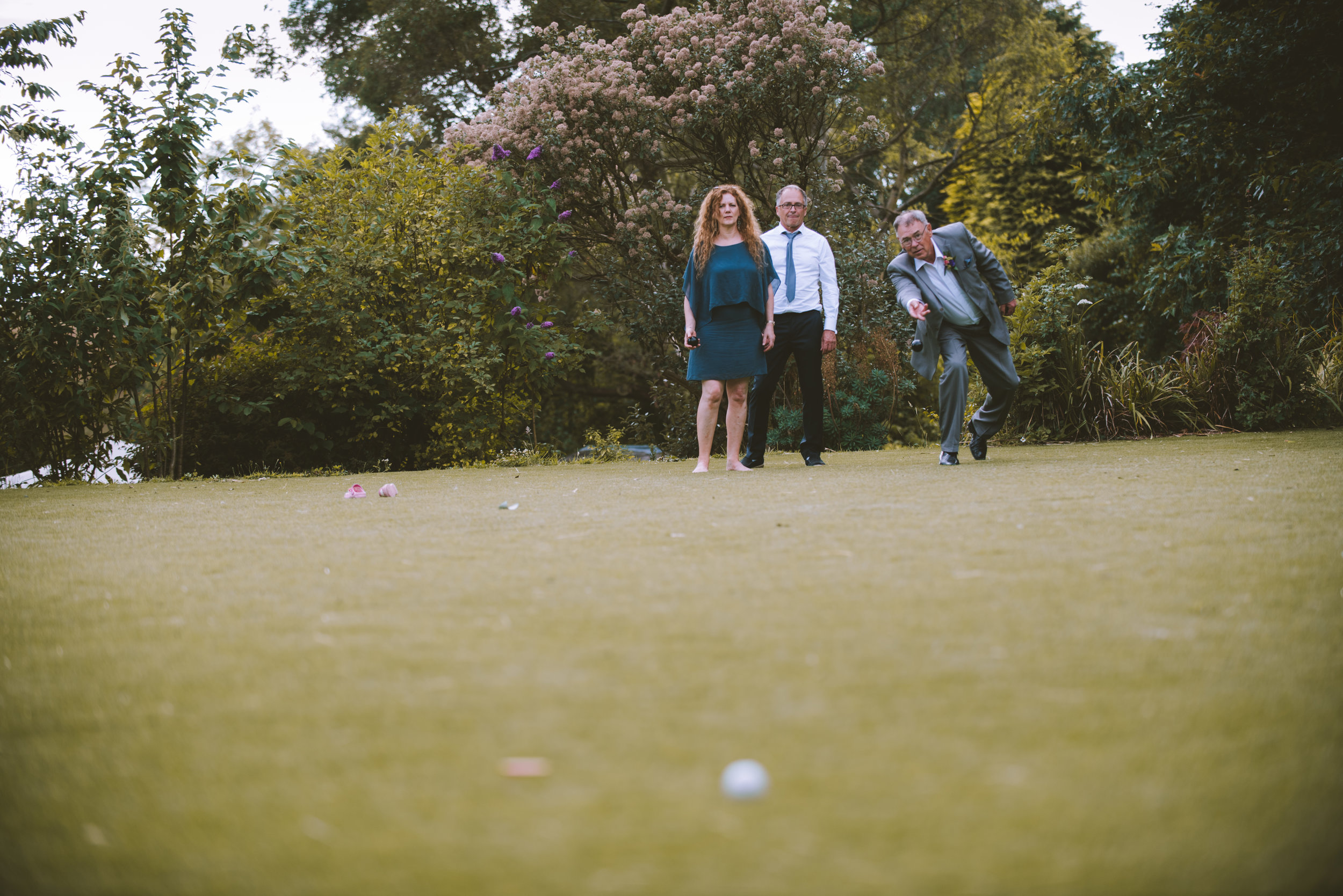 Four people playing a game of bocce on a green lawn in a garden with trees and flowering bushes.