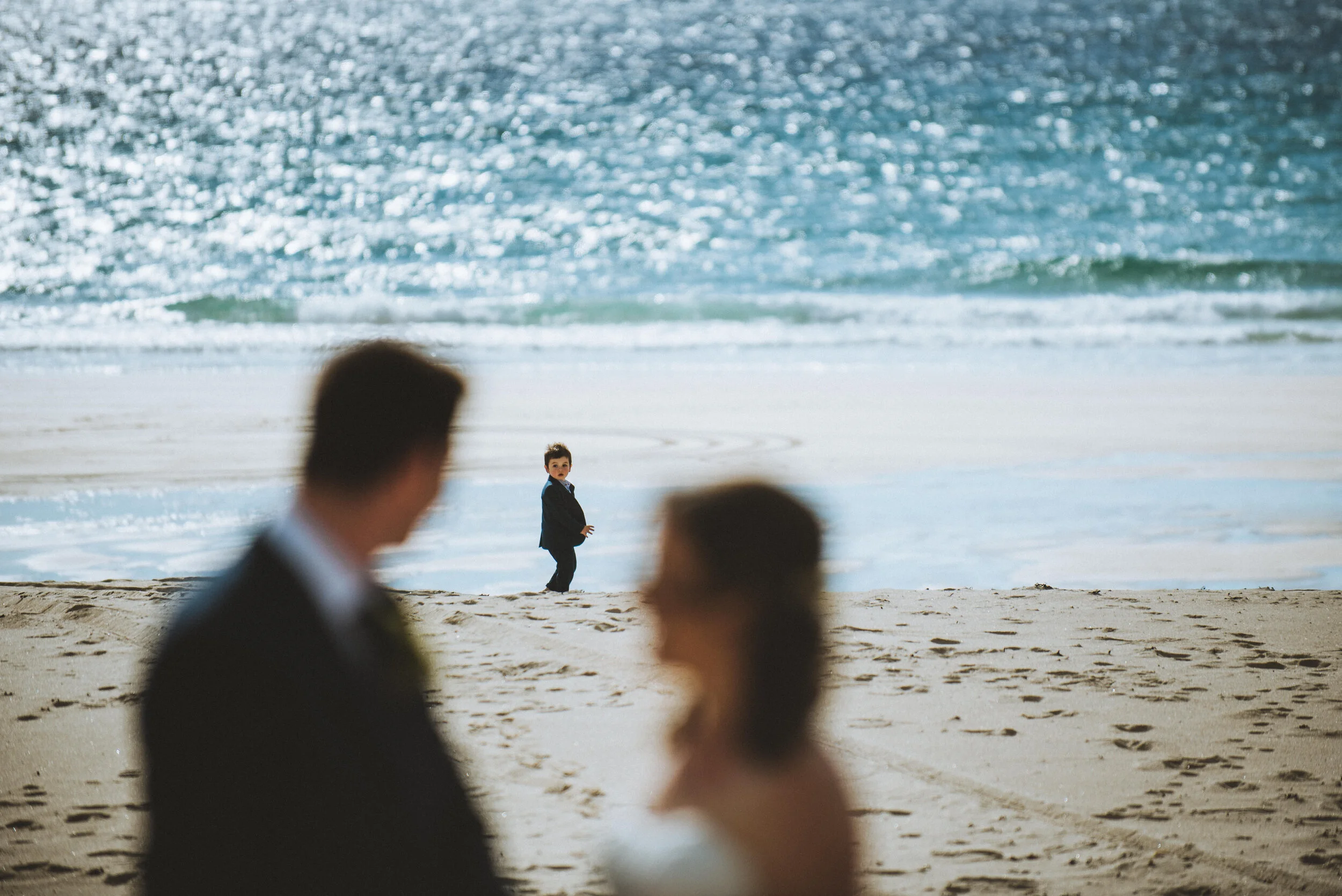 A blurred man and woman in wedding attire in the foreground, with a young boy in a suit standing on the beach near the water in the background.