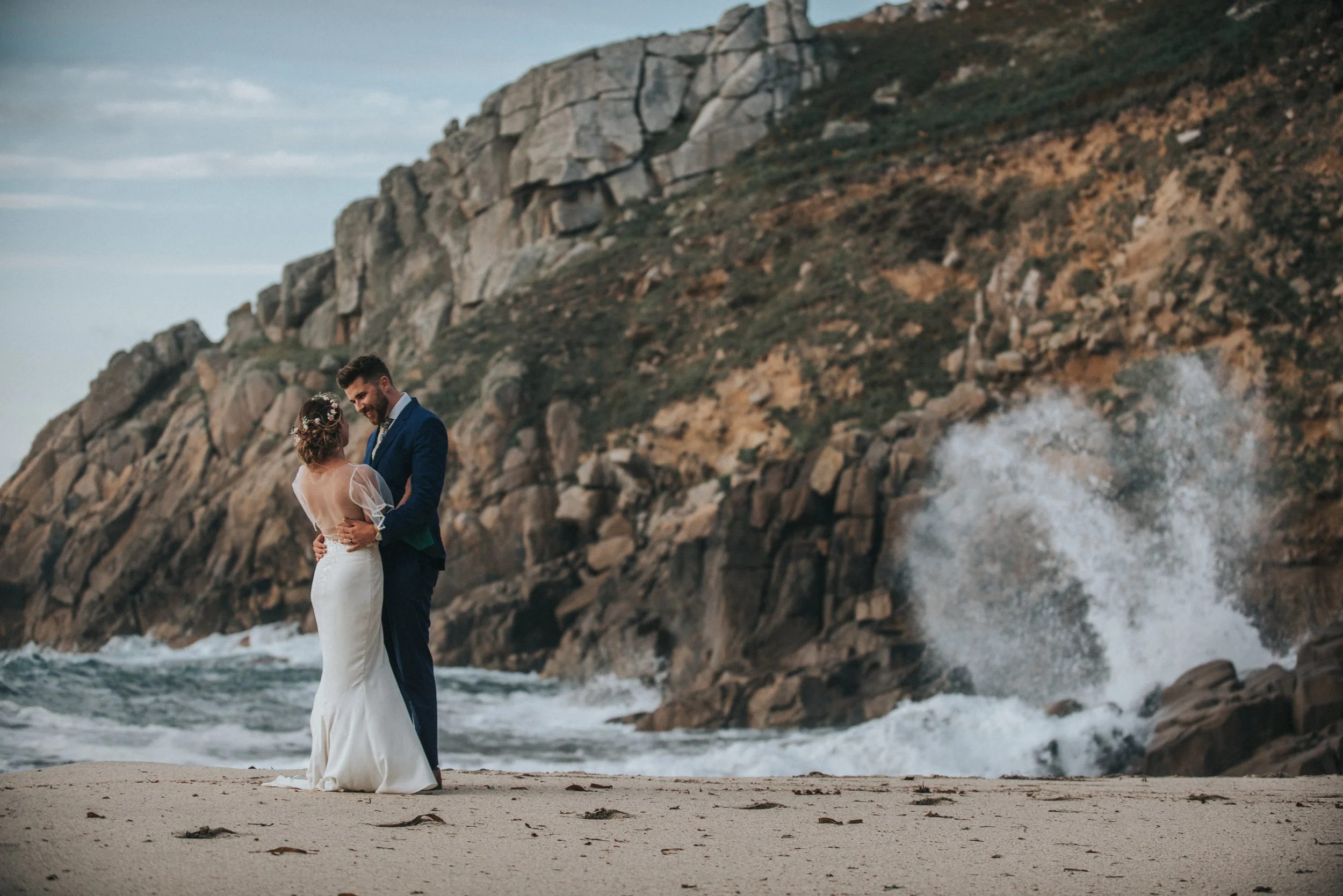 A bride and groom standing on a sandy beach, embracing and smiling at each other with a rocky cliff and ocean waves crashing behind them.