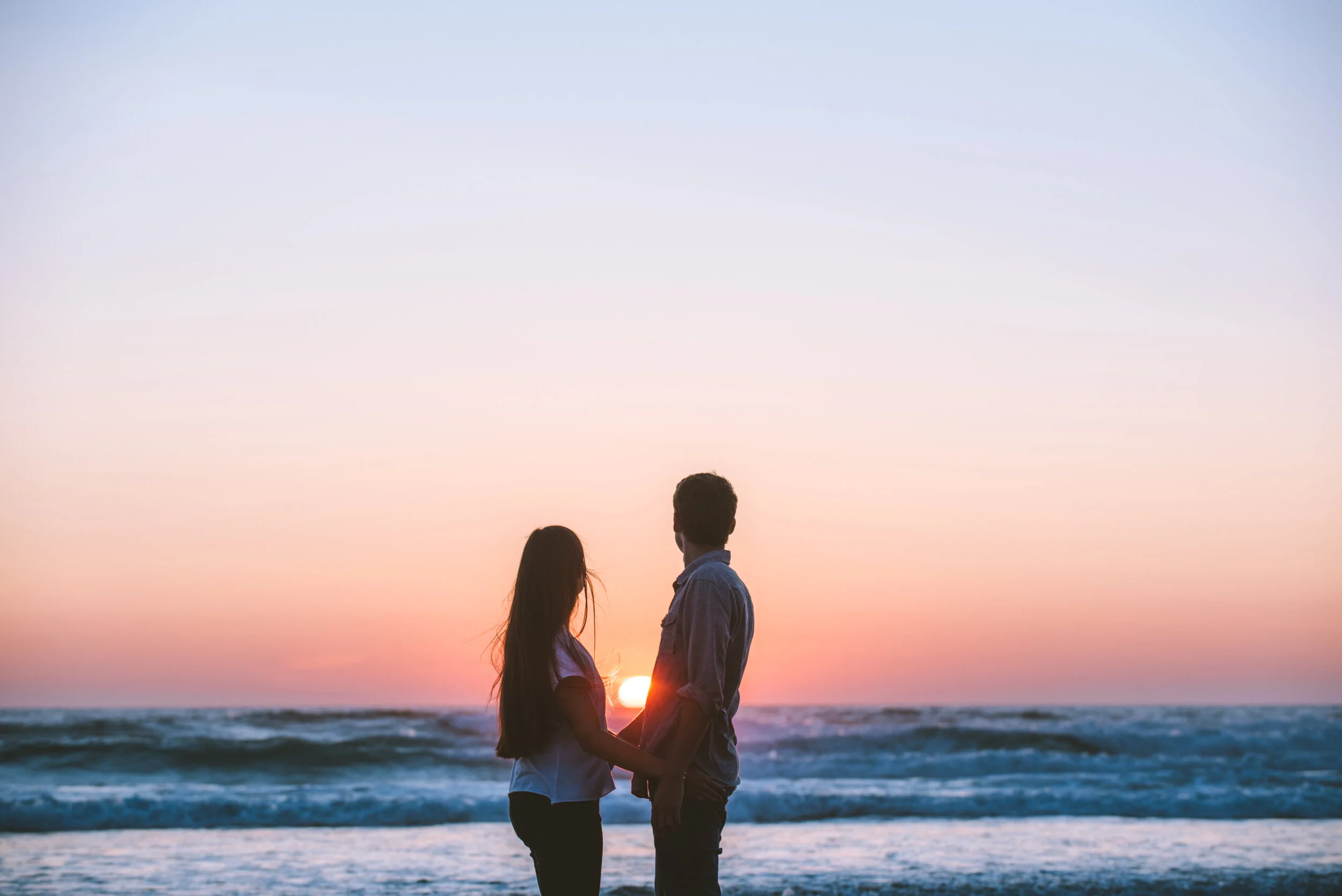 A silhouette of a couple holding hands on the beach during sunset, with the ocean and colorful sky in the background.
