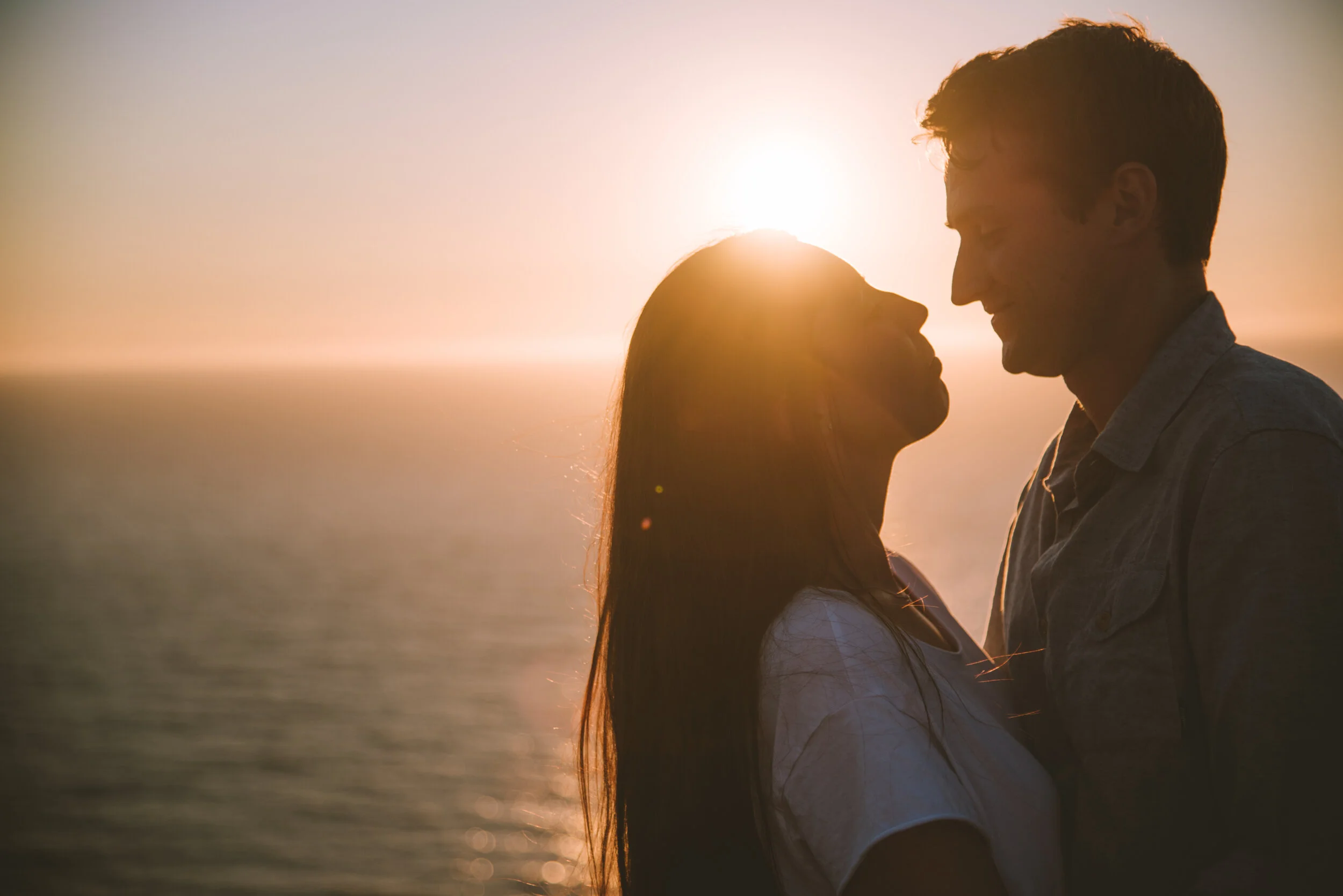 A couple standing close together, facing each other, during sunset over the ocean, with the sun behind them creating a silhouette.