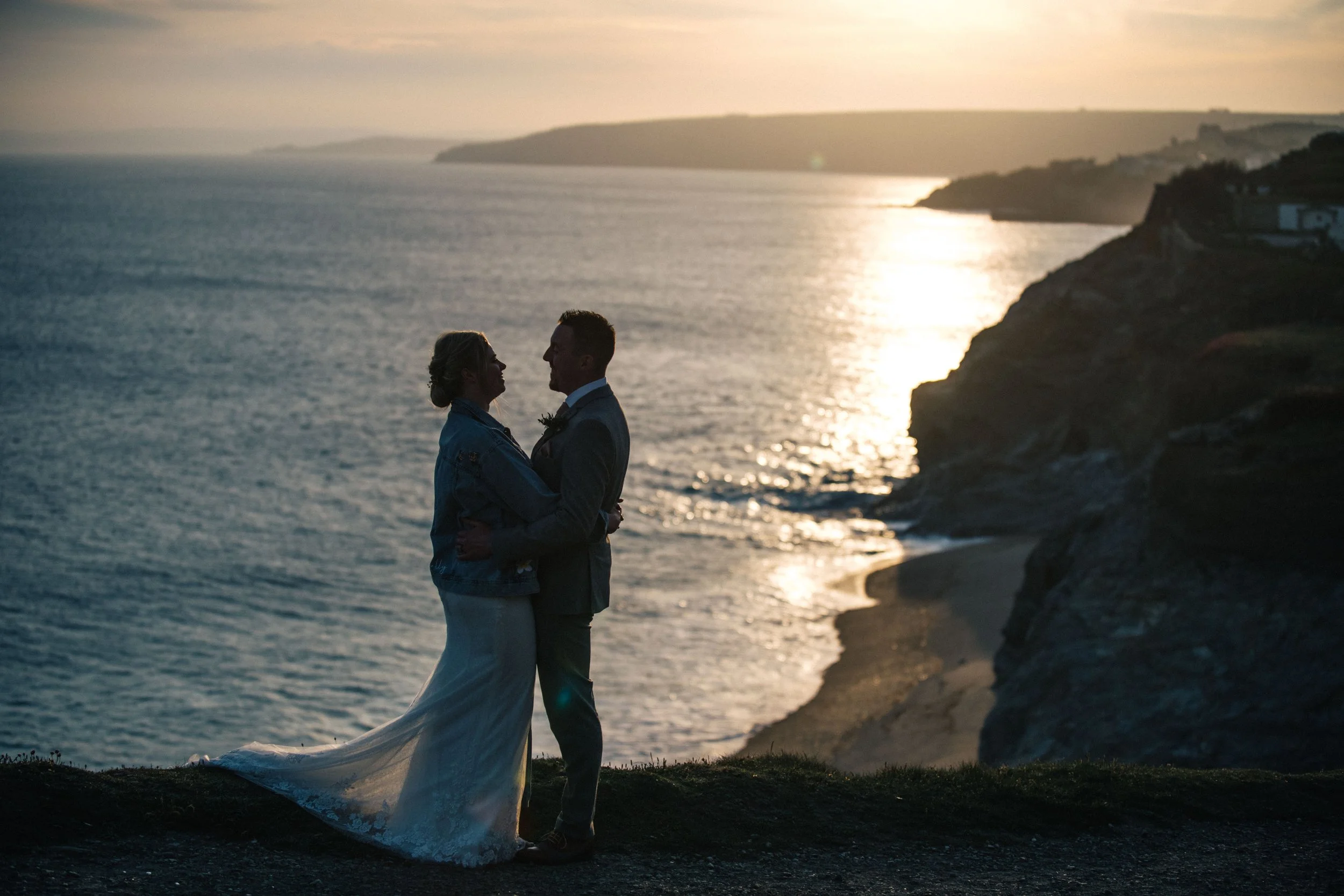 Silhouetted couple standing close together on a cliff at sunset, overlooking the ocean with waves crashing against the rocky shore.