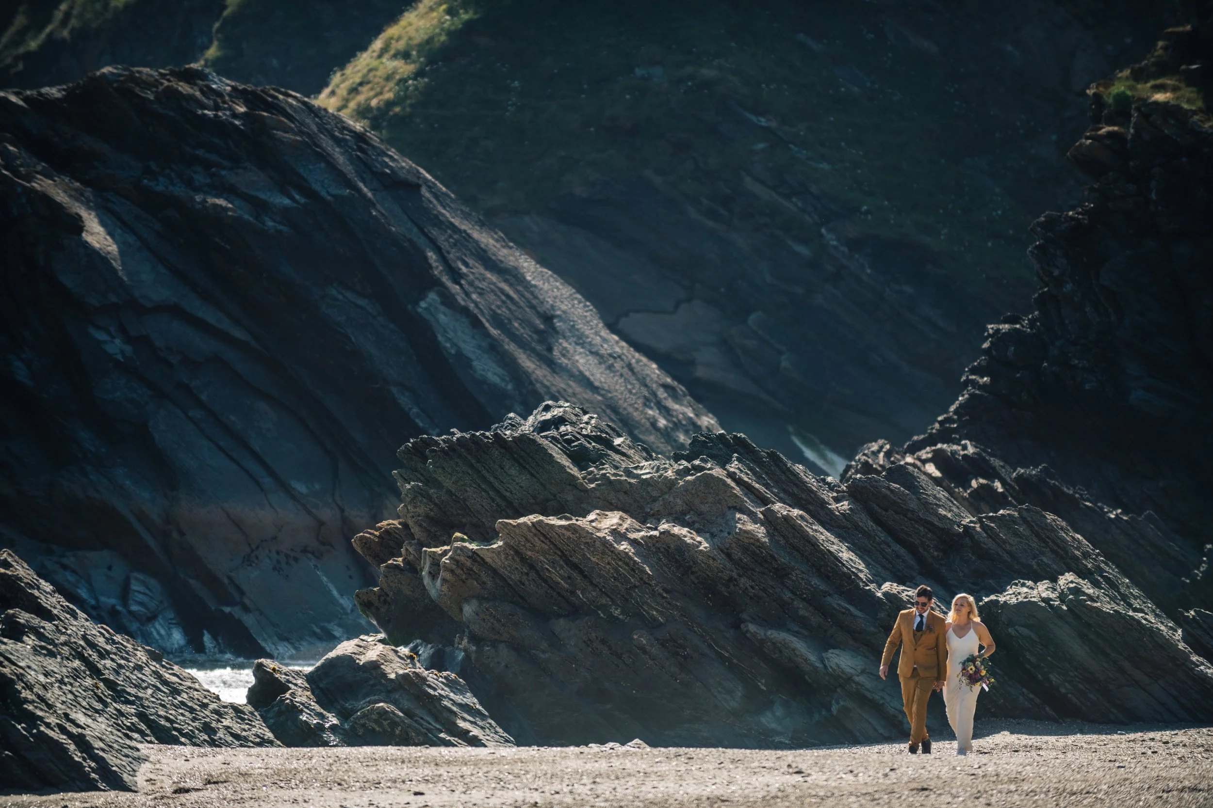 A couple dressed in wedding attire walking along a sandy beach with large dark rocks and cliffs in the background.
