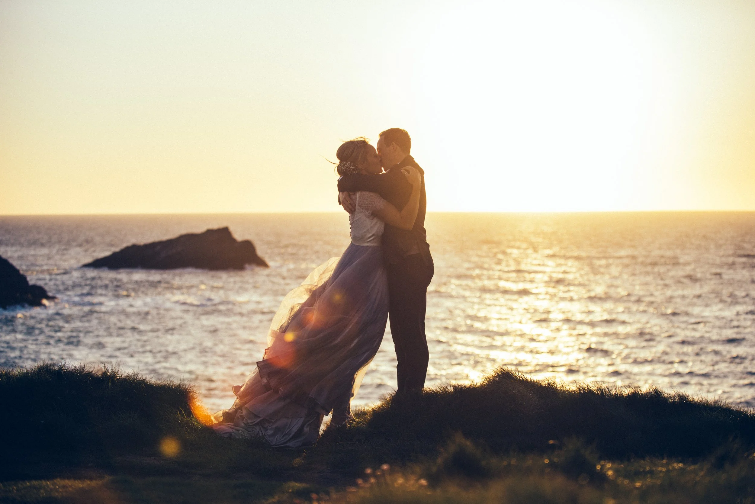A couple kissing on a grassy hill near the ocean during sunset, with rocks in the background.