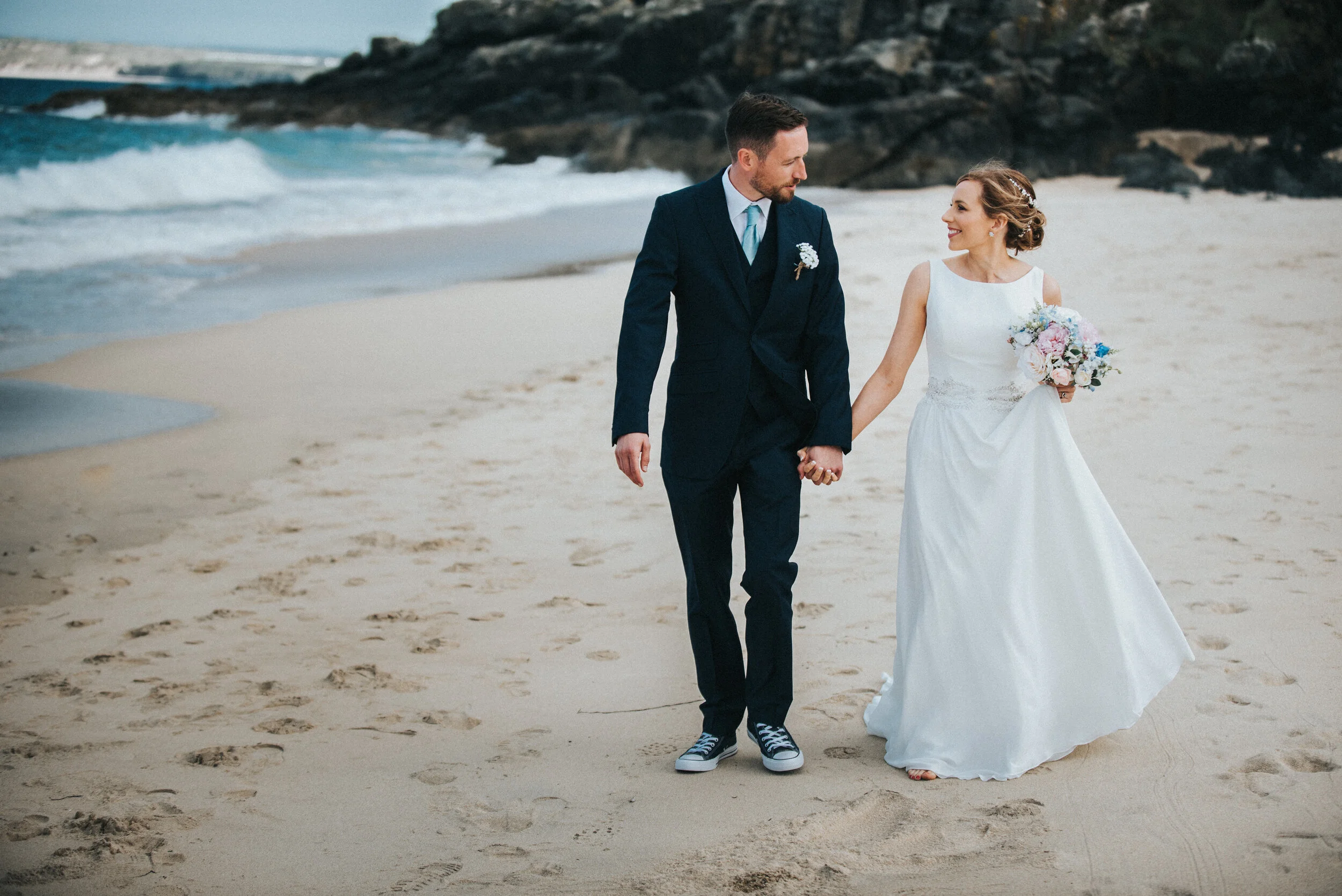 A bride and groom walking on the beach, holding hands, with the ocean and rocks in the background.