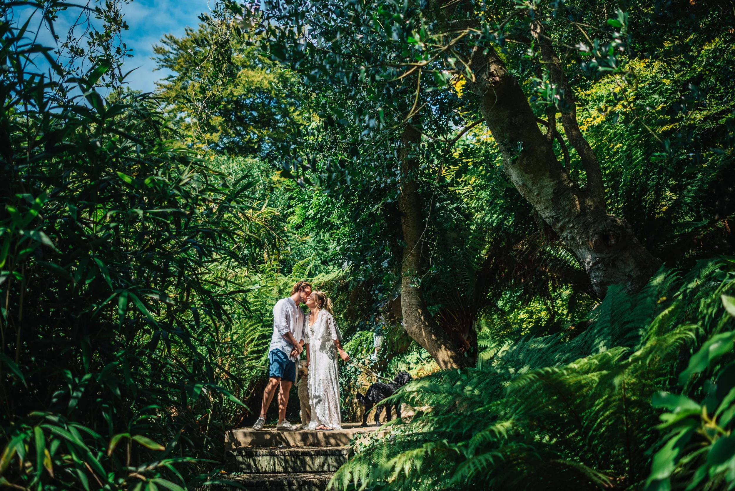 A couple kissing on a stone path surrounded by lush green trees and foliage.