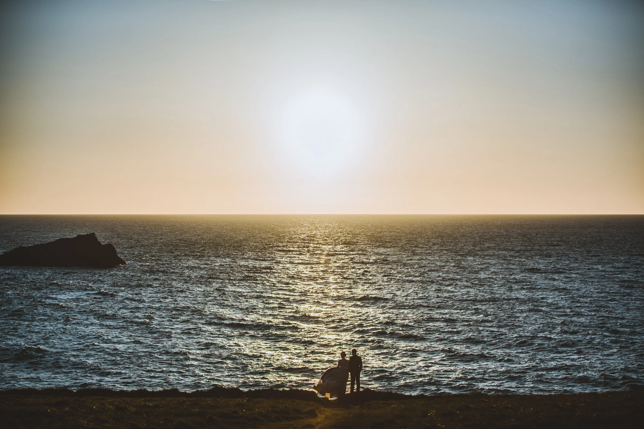 Two people, one sitting and one standing, on a grassy cliff overlooking the ocean at sunset.