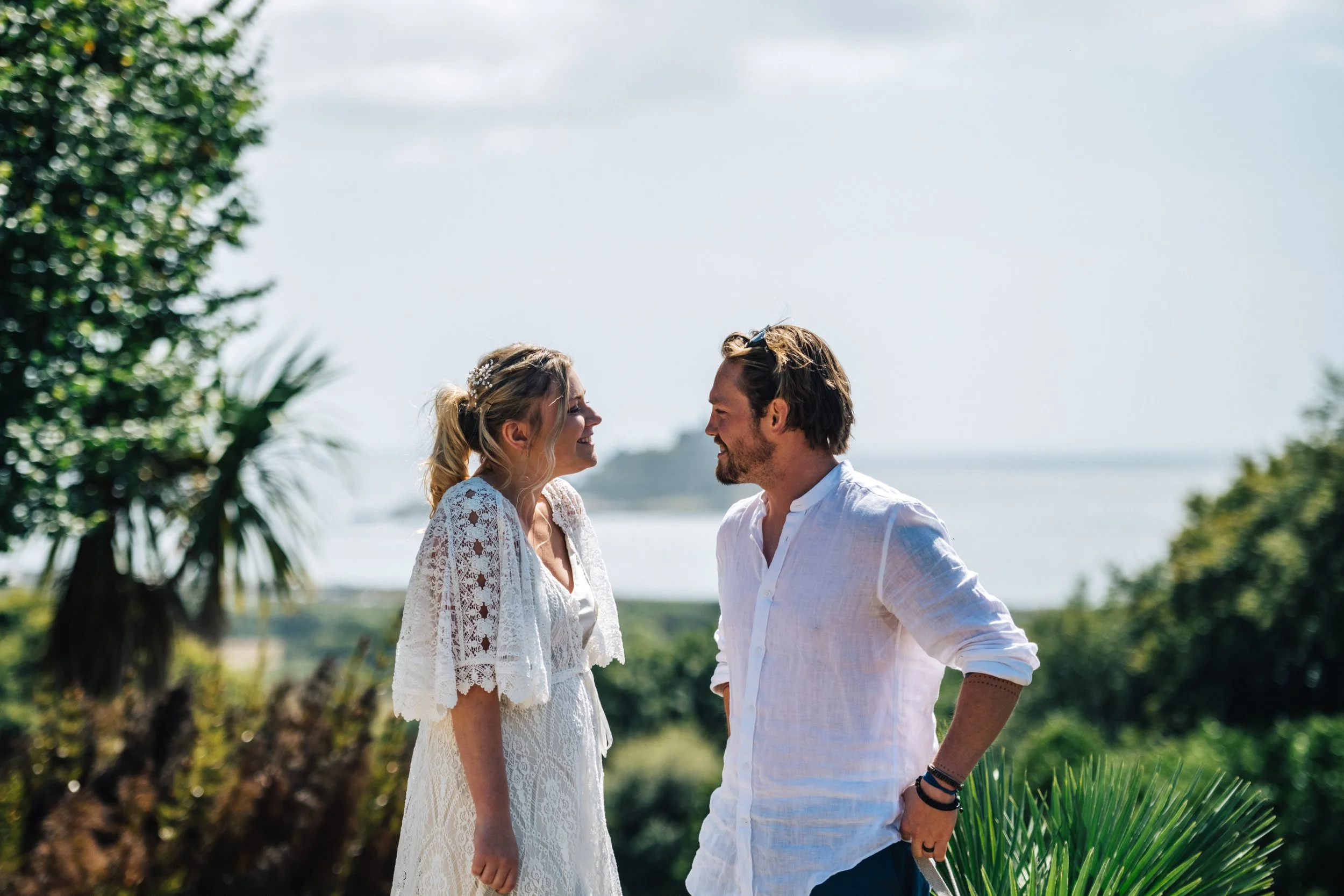 A couple smiling and looking at each other outdoors near a body of water, with greenery and trees in the background.