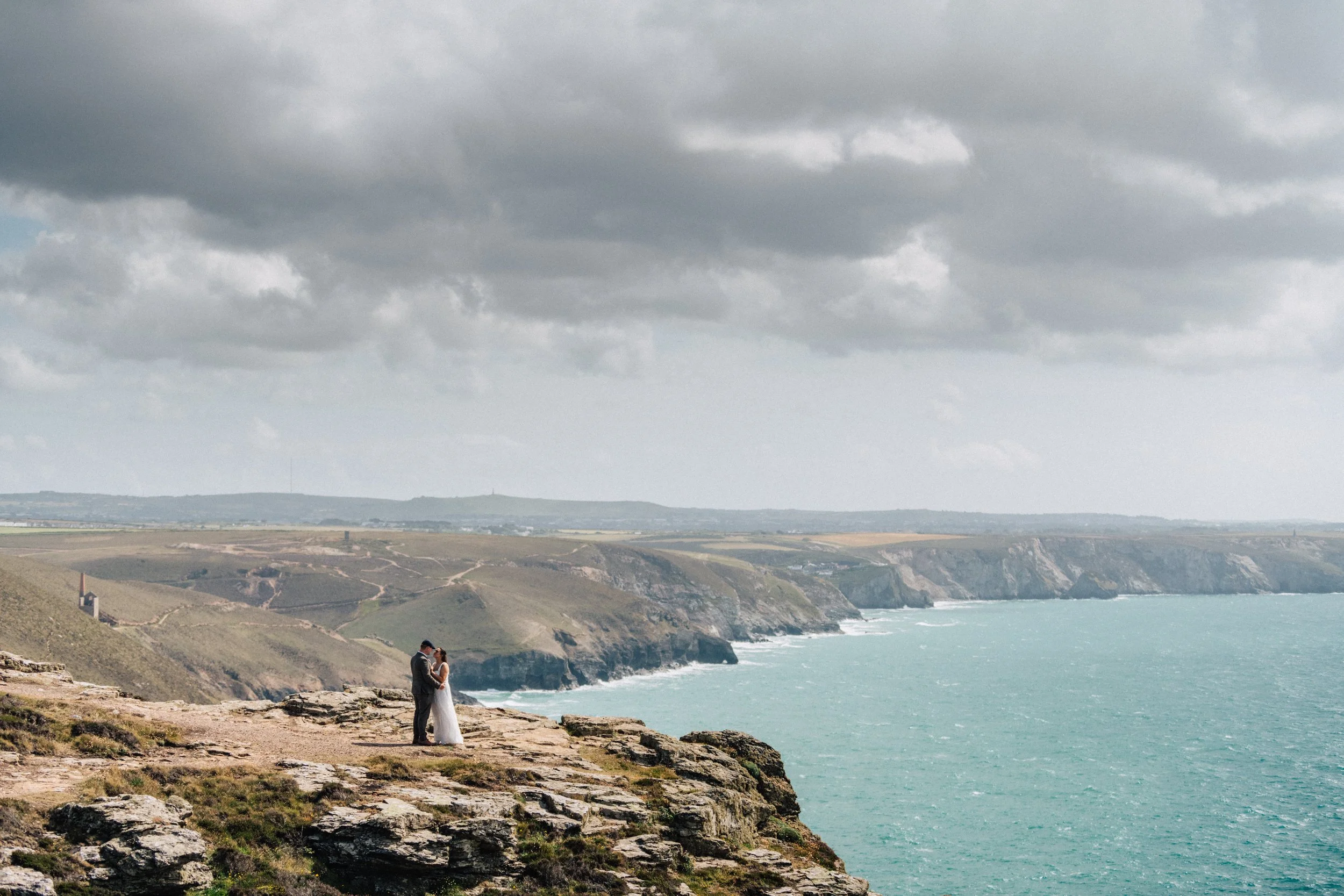 A couple, dressed in wedding attire, standing on a rocky cliff overlooking an ocean with steep cliffs and a cloudy sky.