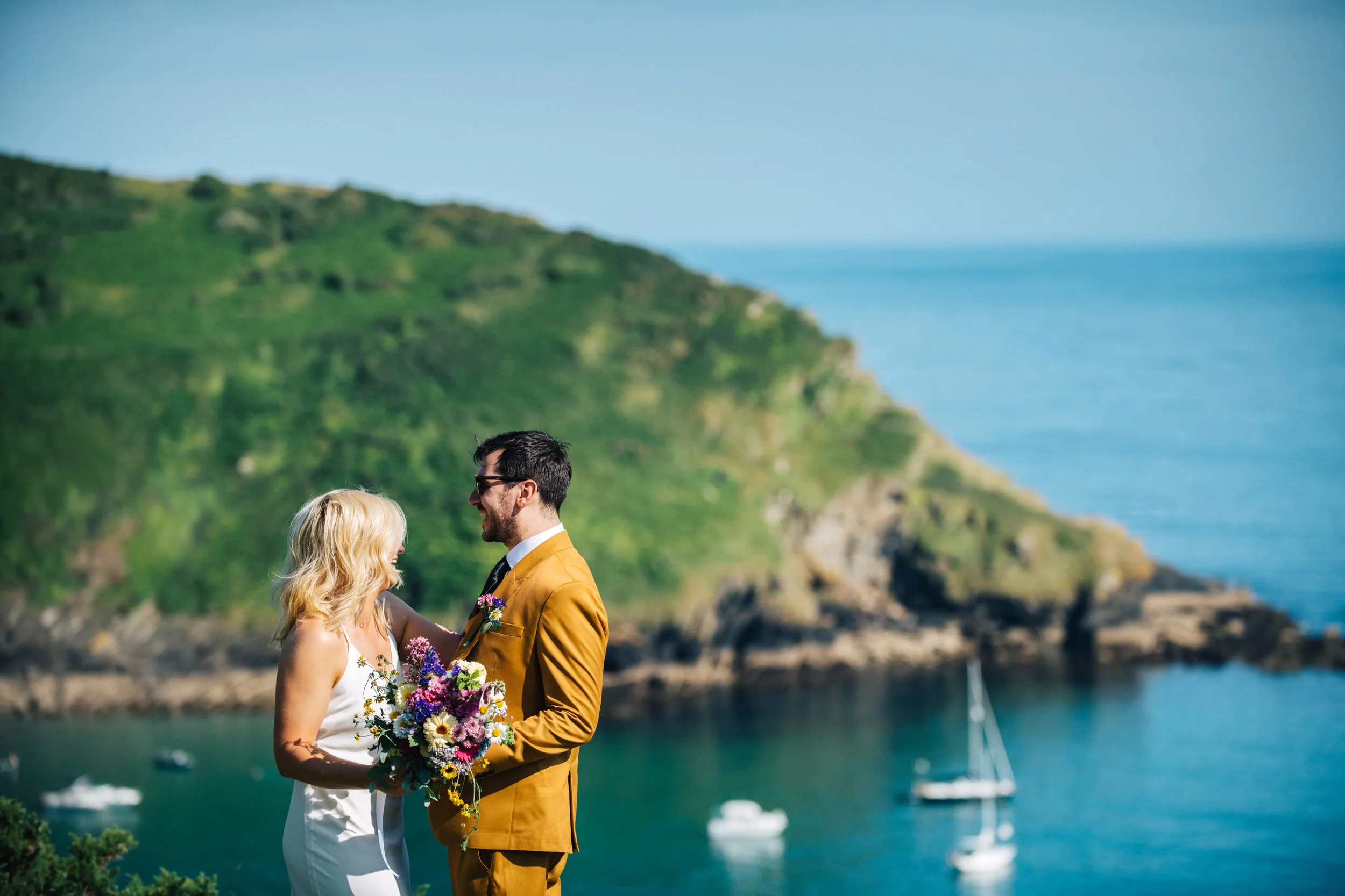 A couple standing on a scenic seaside, with sailboats in the water and green hills in the background, during a wedding or romantic occasion.