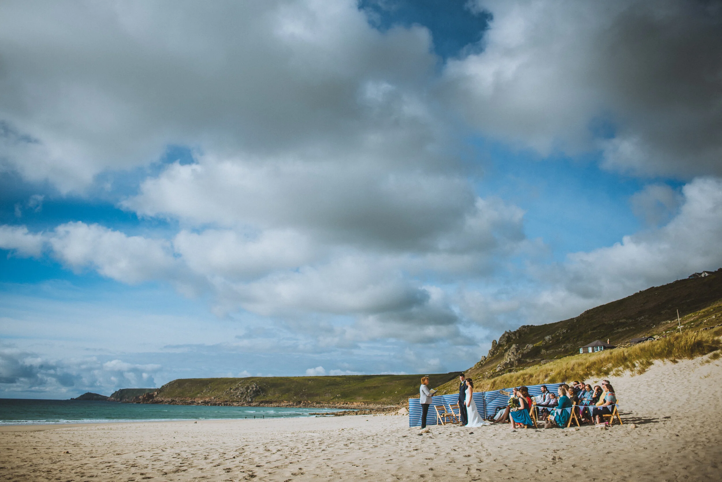 A beach wedding ceremony with guests seated on chairs, a bride and groom standing under a blue and white striped cloth, officiant, on a sandy beach with green hills and cloudy sky in the background.
