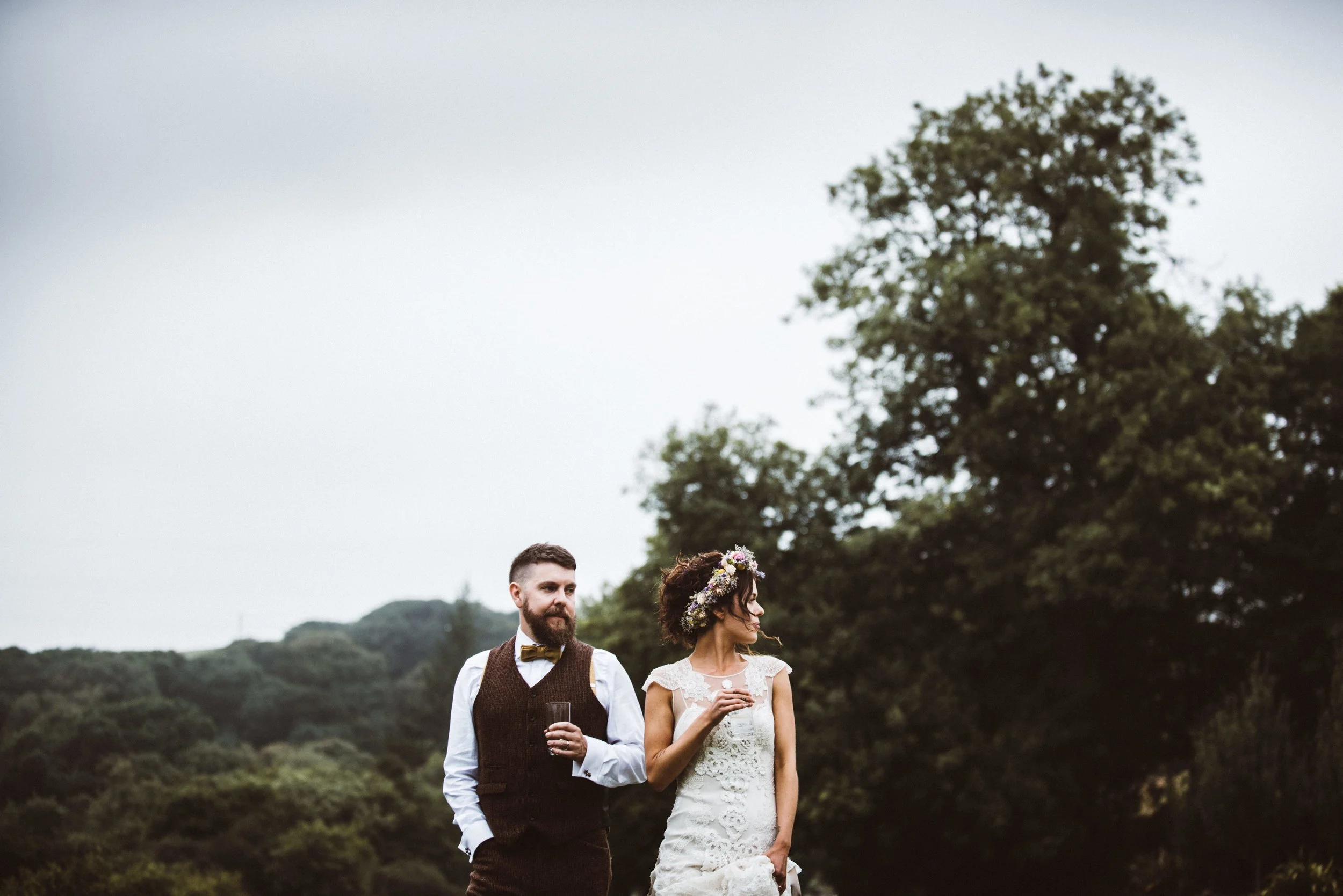 A man and woman in wedding attire standing outdoors, with a backdrop of trees and cloudy sky. The woman is wearing a lace wedding dress and a floral crown, holding a glass, while the man is in a vest, white shirt, and bow tie, holding a glass, both l