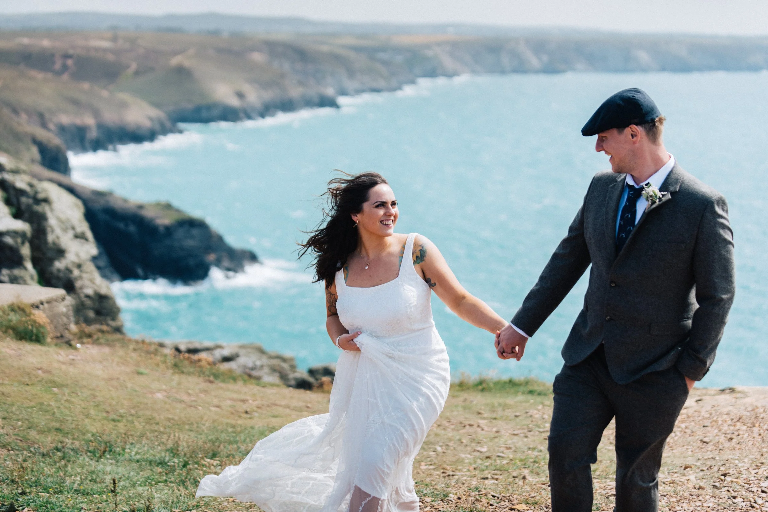 A bride and groom holding hands and smiling on a grassy cliffside overlooking the ocean.