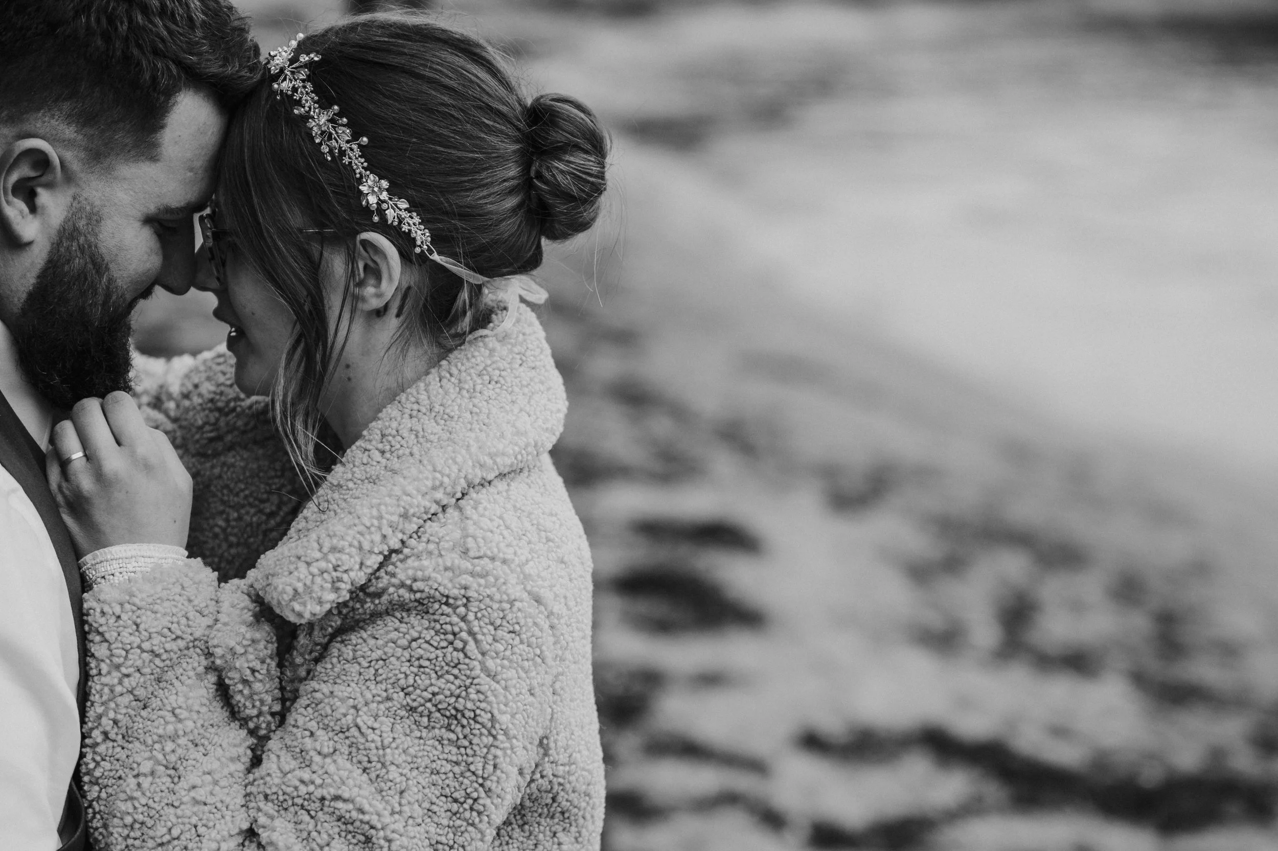 A couple touching foreheads and smiling, standing close to each other by the water, with the woman wearing a floral headpiece and a textured coat, and the man wearing a vest.