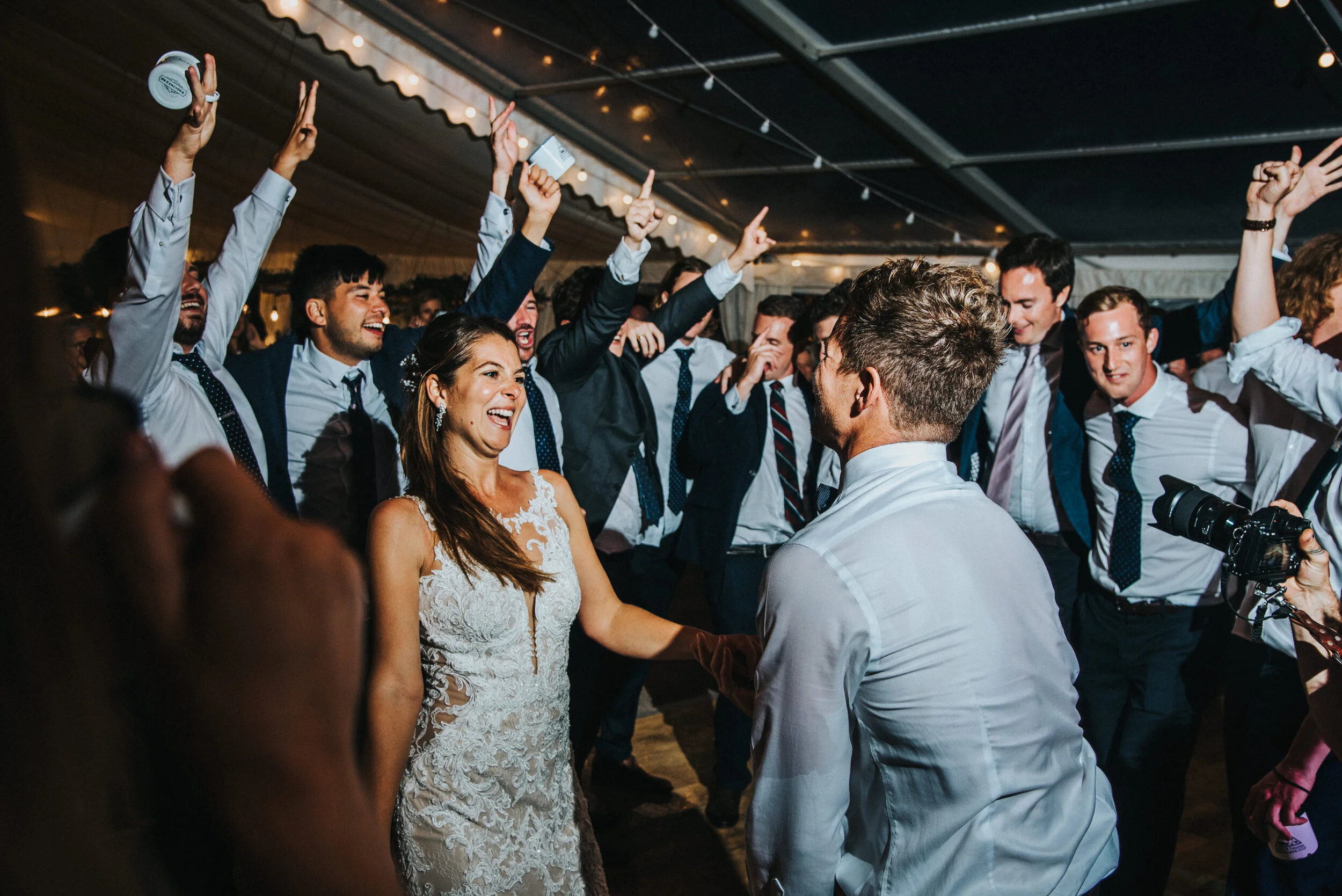 Daymer bay wedding photography captured by Cornwall wedding photographer Mark Shaw Photography -A bride and groom dancing with friends at a wedding reception, with guests raising their hands and smiling under string lights in a decorated tent.