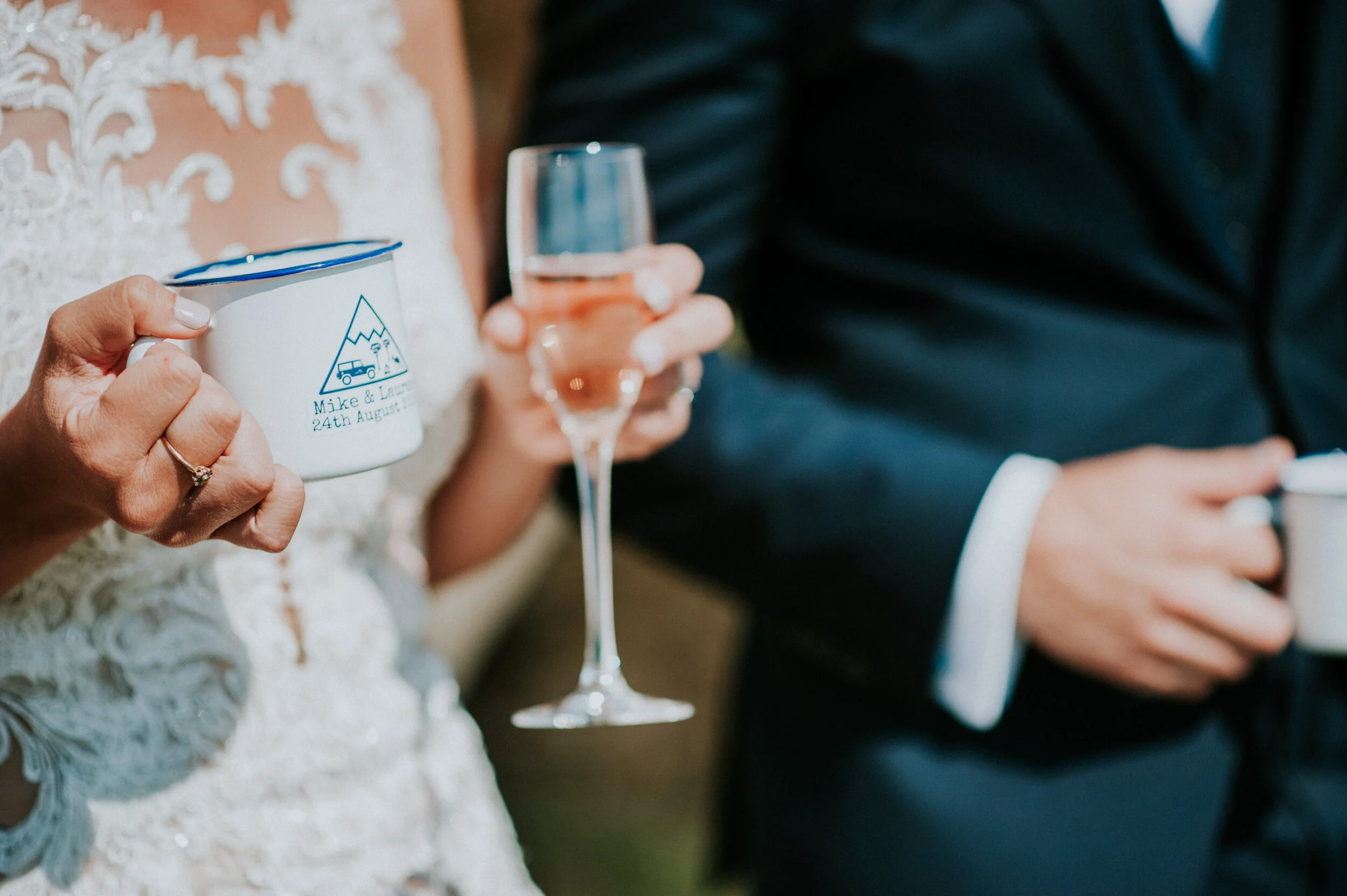 Wedding photo at Greenaway Field in Cornwall. A bride in a lace wedding dress holding a white mug with a mountain and vehicle illustration, and a groom in a dark suit holding a glass of champagne, during a wedding celebration.