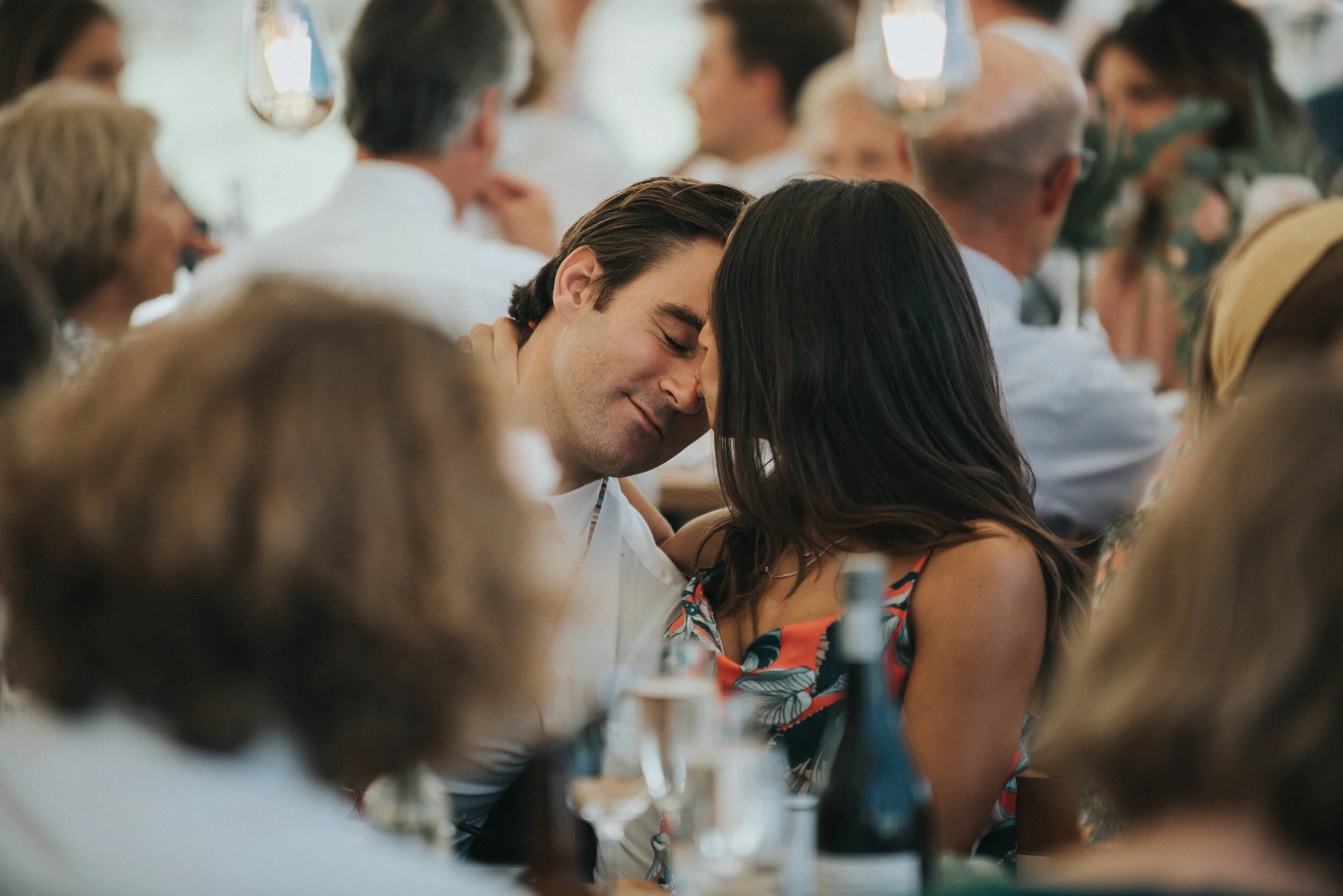 Daymer bay wedding photography in Cornwall captured by Mark Shaw Photography - A romantic moment at a gathering, with a man and woman leaning their foreheads together, smiling softly with eyes closed, surrounded by other people seated at a table.