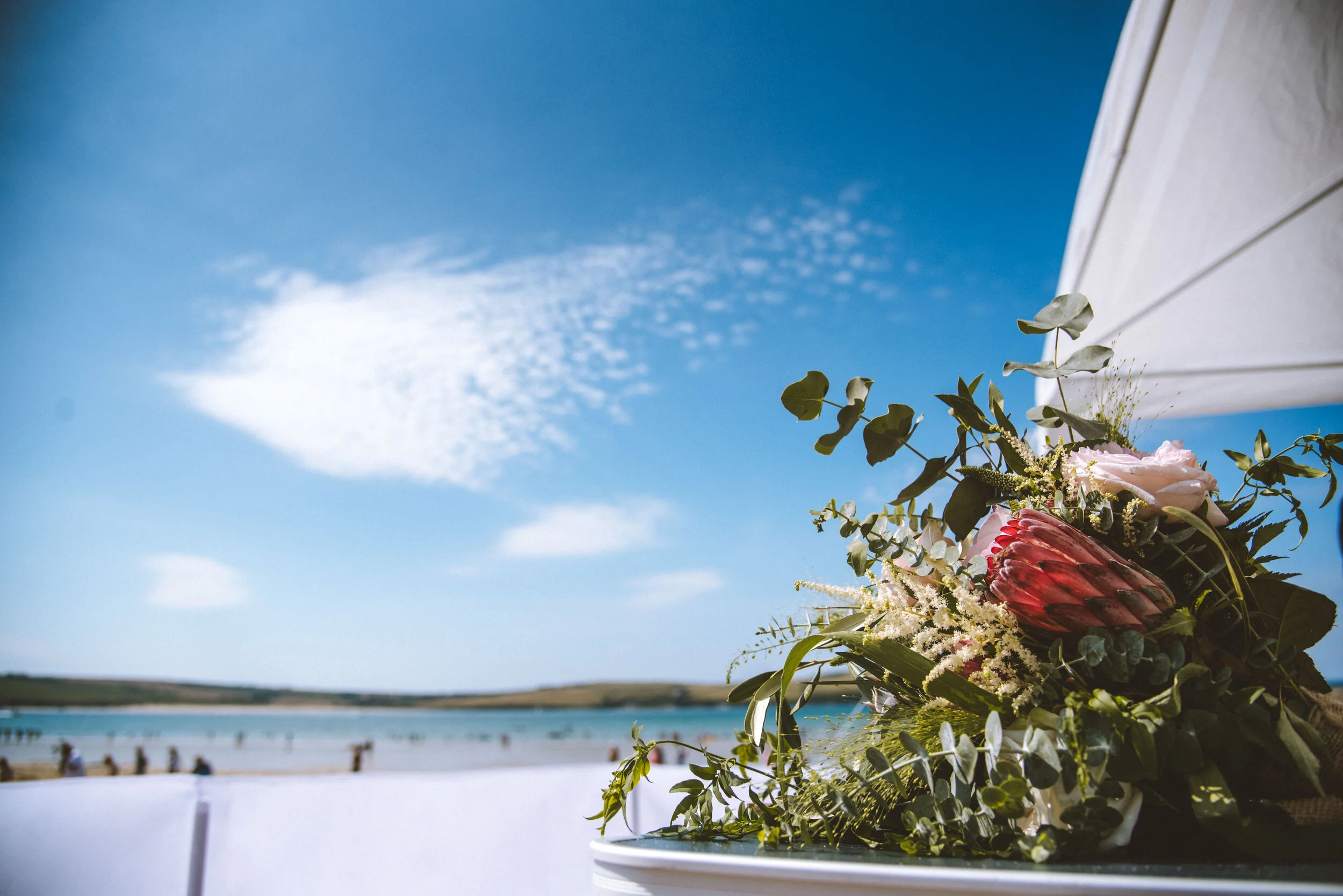 A bouquet of pink and white flowers with greenery on a table outdoors with a beach, water, and blue sky in the background.